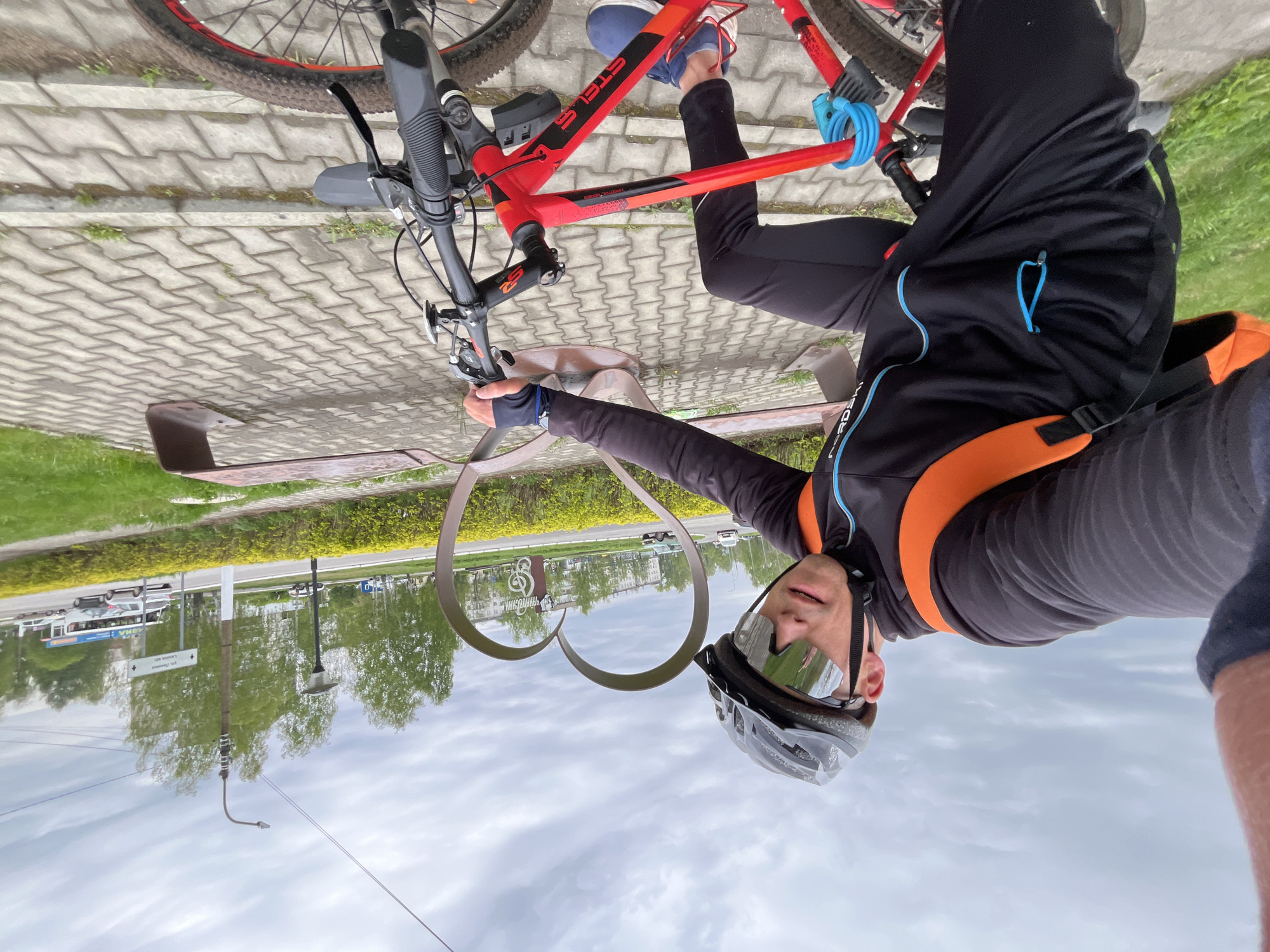 The image shows a person wearing a cycling helmet and mirrored glasses, standing next to a red mountain bike. The bike is secured with a blue cable lock. The person is wearing a dark jacket with orange and blue accents, as well as black cycling pants. The background features a paved area with a bench, a decorative metal heart-shaped structure, and a view of a street with trees and buildings. A signpost is visible in the background with Cyrillic text. Based on the sign "ЧАЙКОВСКИЙ" prominently featured in the metal heart shape and the Cyrillic text on the signpost, the location is identified as the city of Chaikovsky, Russia.