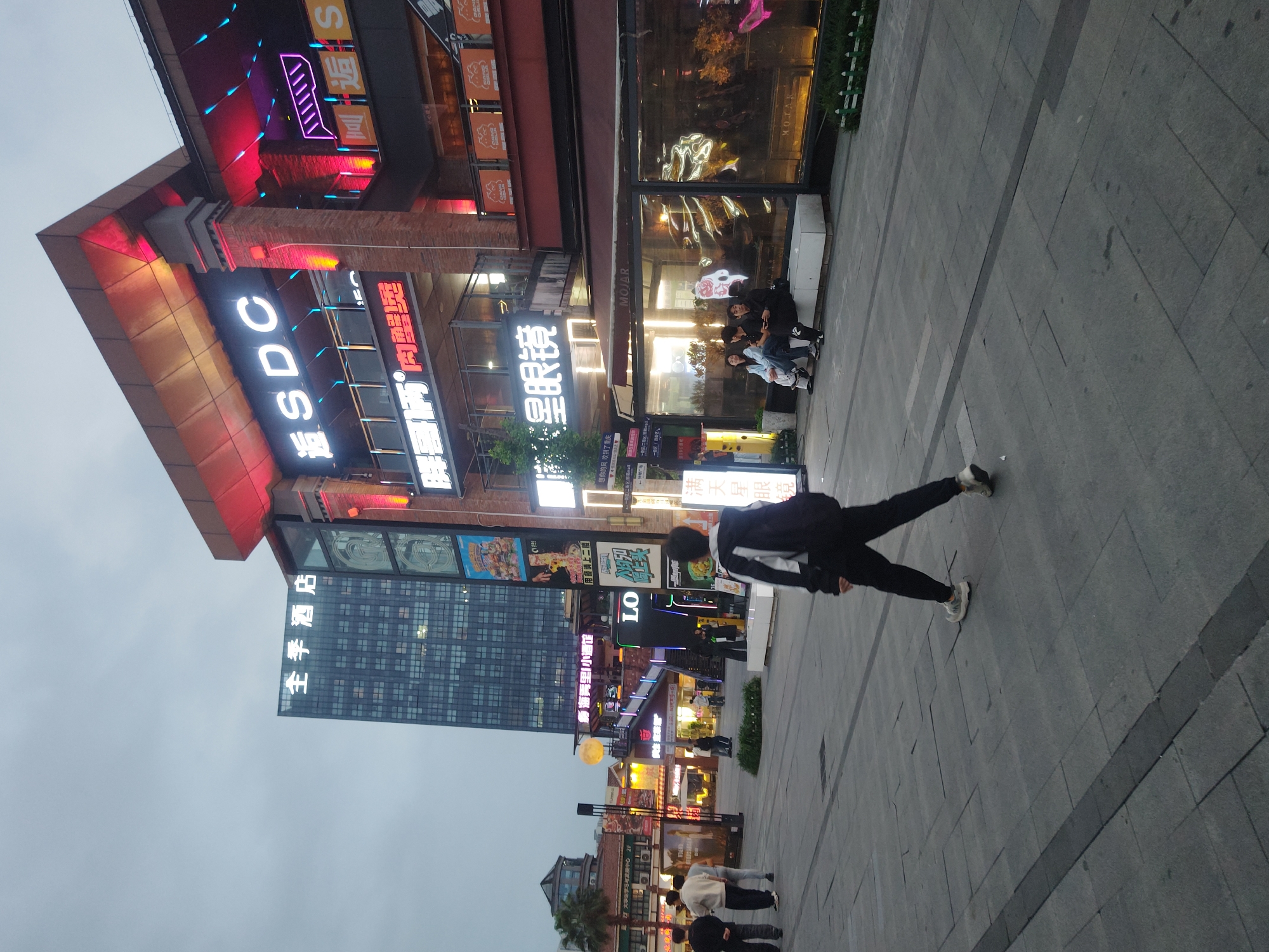 The image captures an evening or dusk scene in an urban commercial area, specifically in the Chongqing High-tech Zone, China. The sky is overcast and light, contrasting with the vibrant illumination of numerous neon signs on the buildings.
The street is a wide pedestrian walkway made of grey and dark grey paving stones. On the left, a multi-story building features a distinct reddish-orange angled roof and brick elements. Its facade is adorned with multiple brightly lit signs, including "逅SDC" in large white characters and "大学生学习考就服中心" (University Student Study and Examination Service Center). Another sign reads "胖哥两肉蟹煲" (Fat Brother Two Crab Pot) in red text. Further down, another building has "全季酒店" (Ji Hotel) in white text.
On the right side of the street, more modern buildings with large glass windows house various businesses. One prominent sign features the word "MOJAR" in white letters, appearing on an awning and a storefront. Below, a group of people are seated casually outside a building, possibly a cafe or restaurant. Another sign nearby says "星眼镜" (Star Eyeglasses). Further down the street, more illuminated signs and storefronts are visible, including one with "满天星眼镜" (Gypsophila Eyeglasses / Full of Stars Eyeglasses) and a poetic phrase "想你的风吹到了重庆" (The wind that misses you has blown to Chongqing).
In the immediate foreground, a person wearing a dark jacket and trousers is walking away from the camera, stretching or in mid-stride. Other people are scattered along the pedestrian area, suggesting a lively evening atmosphere in this commercial district. The scene is rich with modern urban architecture and glowing commercial signs.
