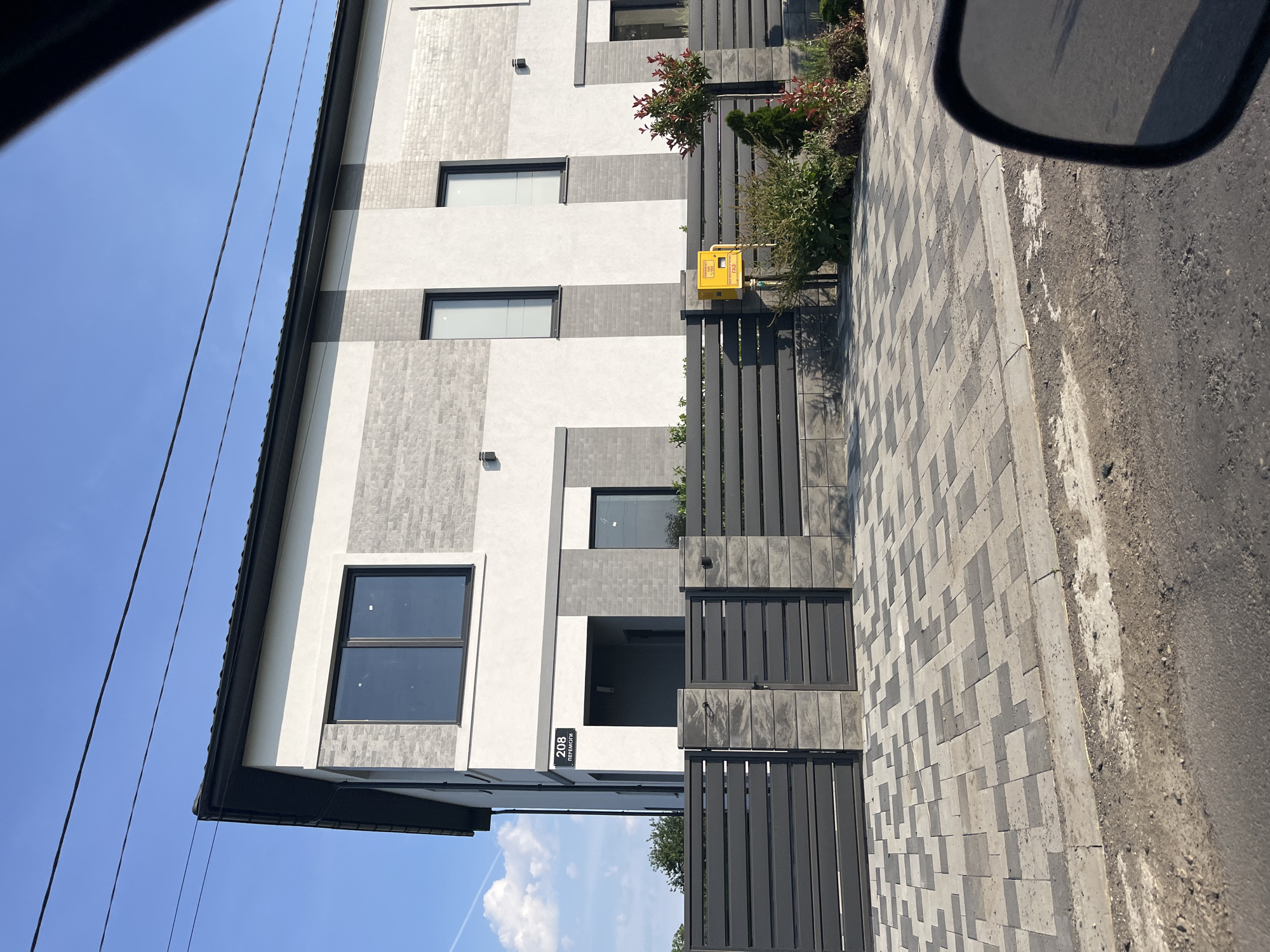 The image shows a modern, two-story residential building with a light gray and white facade, accented with darker gray brickwork. The building features several windows with dark frames. A dark gray horizontal slatted fence with matching gates encloses the property, supported by pillars constructed from gray stone blocks. A paved driveway, laid with interlocking gray and charcoal pavers, leads up to the gates. A yellow utility box, likely for gas, is visible behind the fence, along with some landscaping including a small tree and various shrubs. A street address sign, "208 ПЕРЕМОГИ," is mounted on the building near the entrance. The scene appears to be a residential street in Storozhnytsia, Ukraine, under a clear blue sky with some wispy clouds. Power lines are visible in the upper portion of the image. The perspective suggests the photo was taken from a vehicle.