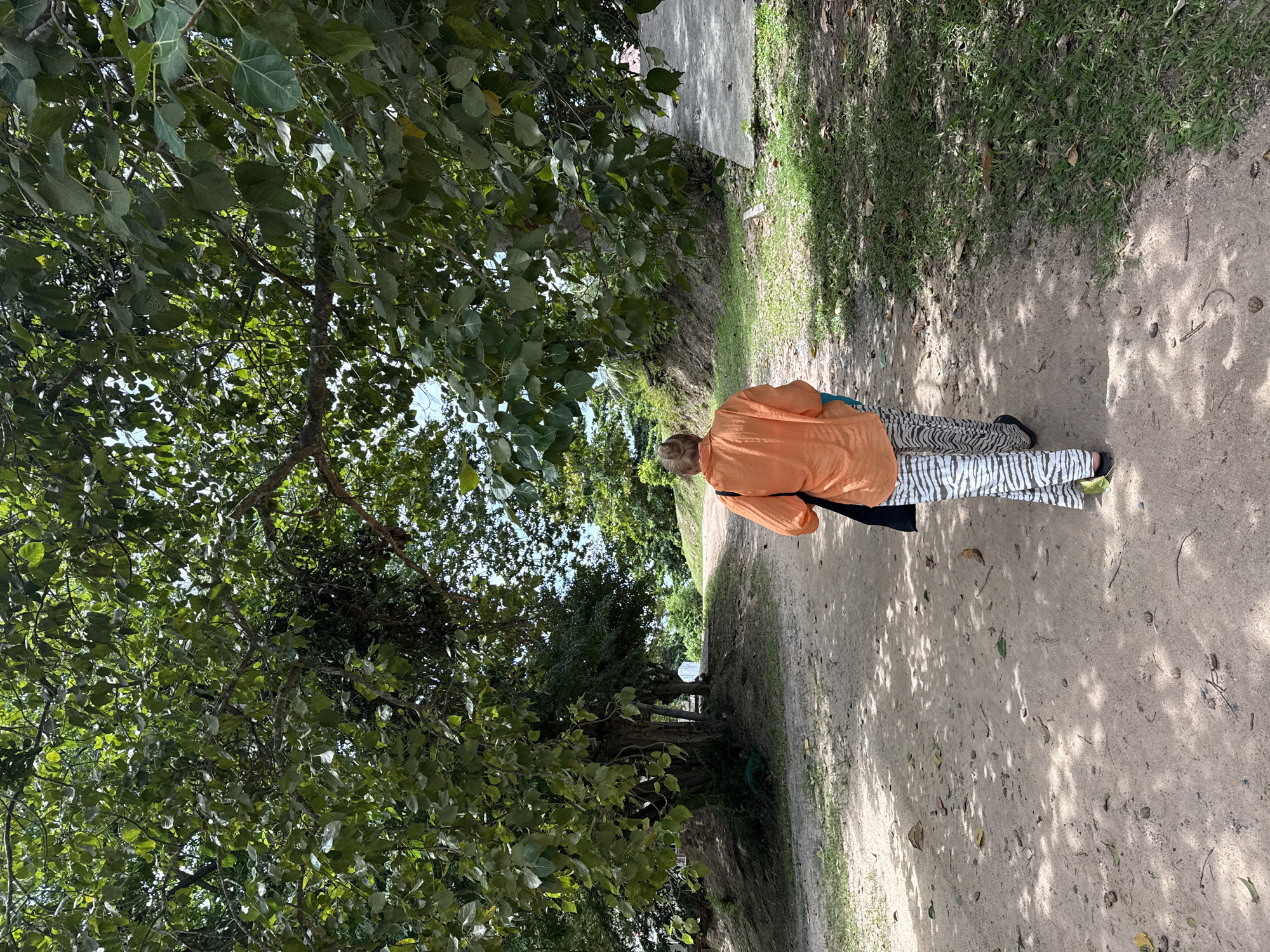 A person wearing an orange shirt and patterned pants walks away from the camera down a dirt path. The path is lined with green grass and trees. The person is carrying a black bag over their shoulder. The scene appears to be in a tropical environment, possibly a park or a residential area. It is daytime and the sun is casting shadows on the ground. The location is Ko Samui District, Thailand.