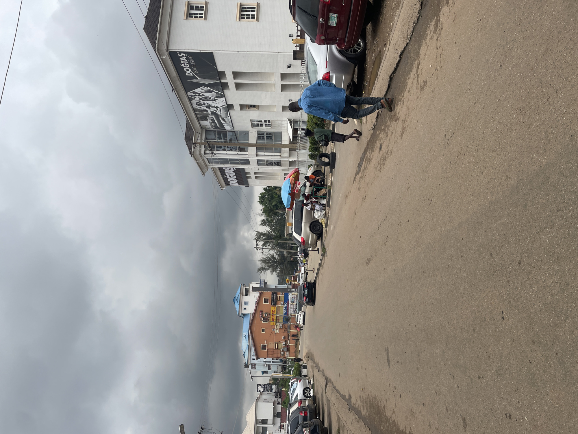 This is a street scene in Abuja, Nigeria, likely on an overcast or cloudy day. The road is paved with asphalt and shows signs of wear. On the right side of the road, a white building with "DOĞTAŞ exclusive" signage is prominently visible. Adjacent to this building, a red SUV is parked. Further down the street, on the left, a colorful building with a blue roof stands out, displaying signs for "DHL" and "TECNO." Several cars are parked along the left side of the street.
In the foreground, two individuals are walking away from the camera. One is wearing a blue shirt and jeans, and the other, a younger person, is in a green shirt and dark shorts. Near the white building, there are a few people gathered around a stall with a blue umbrella, possibly vendors or customers. The environment suggests an urban or commercial area with a mix of modern architecture and more utilitarian buildings. Overhead, power lines are visible against the cloudy sky.