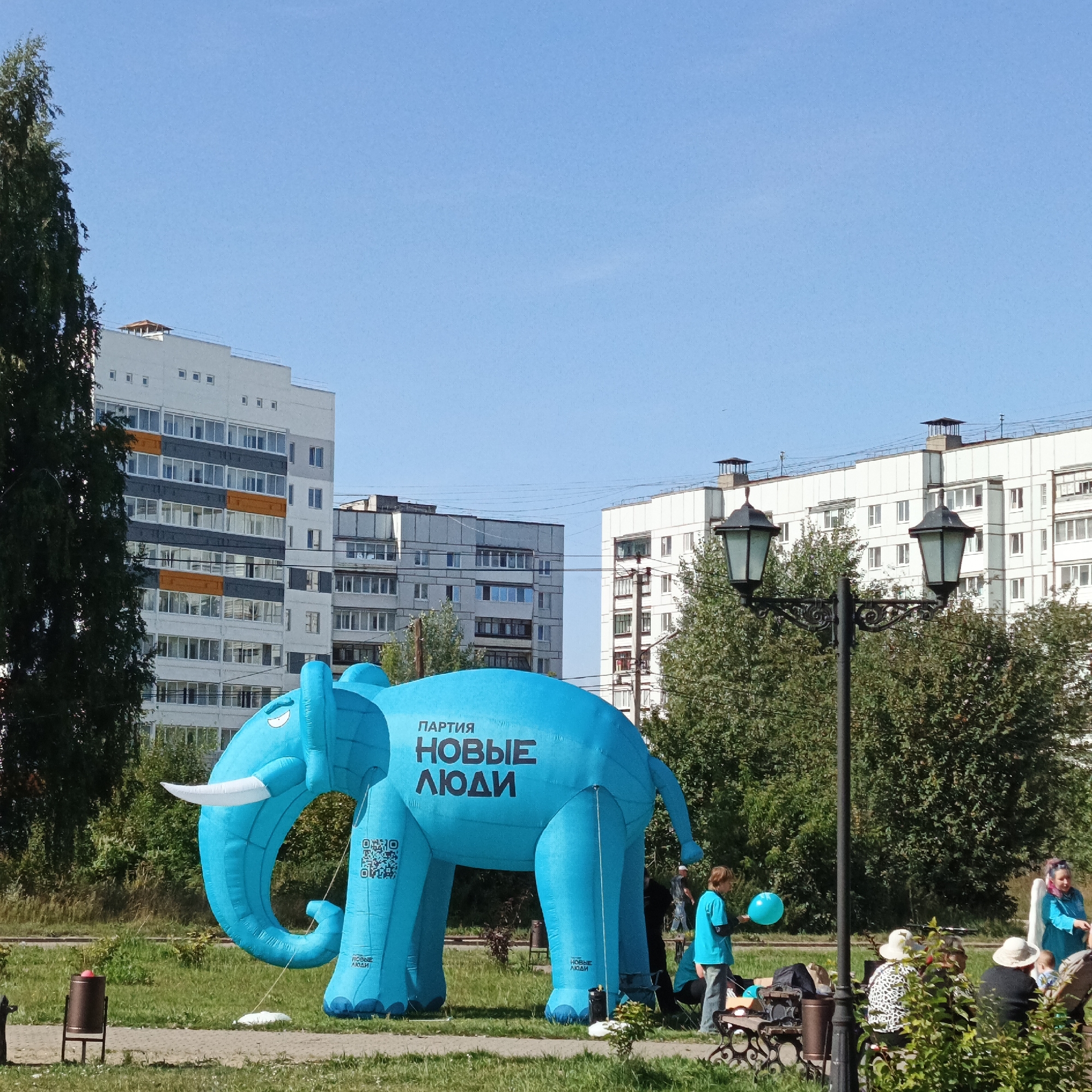 A bright blue inflatable elephant, advertising the "New People Party" (ПАРТИЯ НОВЫЕ ЛЮДИ), is the central feature of this outdoor scene. The elephant is positioned on a grassy area with a paved path in the foreground. Behind the elephant, several modern apartment buildings, typical of the city of Chaikovsky, Russia, rise against a clear blue sky. The buildings are multi-story and feature a mix of white and gray facades with visible windows. Trees and bushes are scattered throughout the park-like setting, with a dark green evergreen tree on the left and deciduous trees and bushes to the right.
Several people are visible in the scene. A young person in a blue t-shirt and jeans holds a blue balloon near the elephant's rear. Further to the right, near some park benches, a group of people are gathered, some seated and others standing. One woman in a blue top with pink-streaked hair appears to be interacting with a baby. Another woman in a white hat and a patterned top is seated on a bench. A trash can is located to the left of the elephant, near the paved path. Two ornate streetlights with traditional lanterns are positioned in the middle ground, adding a classic touch to the otherwise modern urban landscape. The overall impression is a sunny day in a residential area, possibly during a public event or promotional activity.