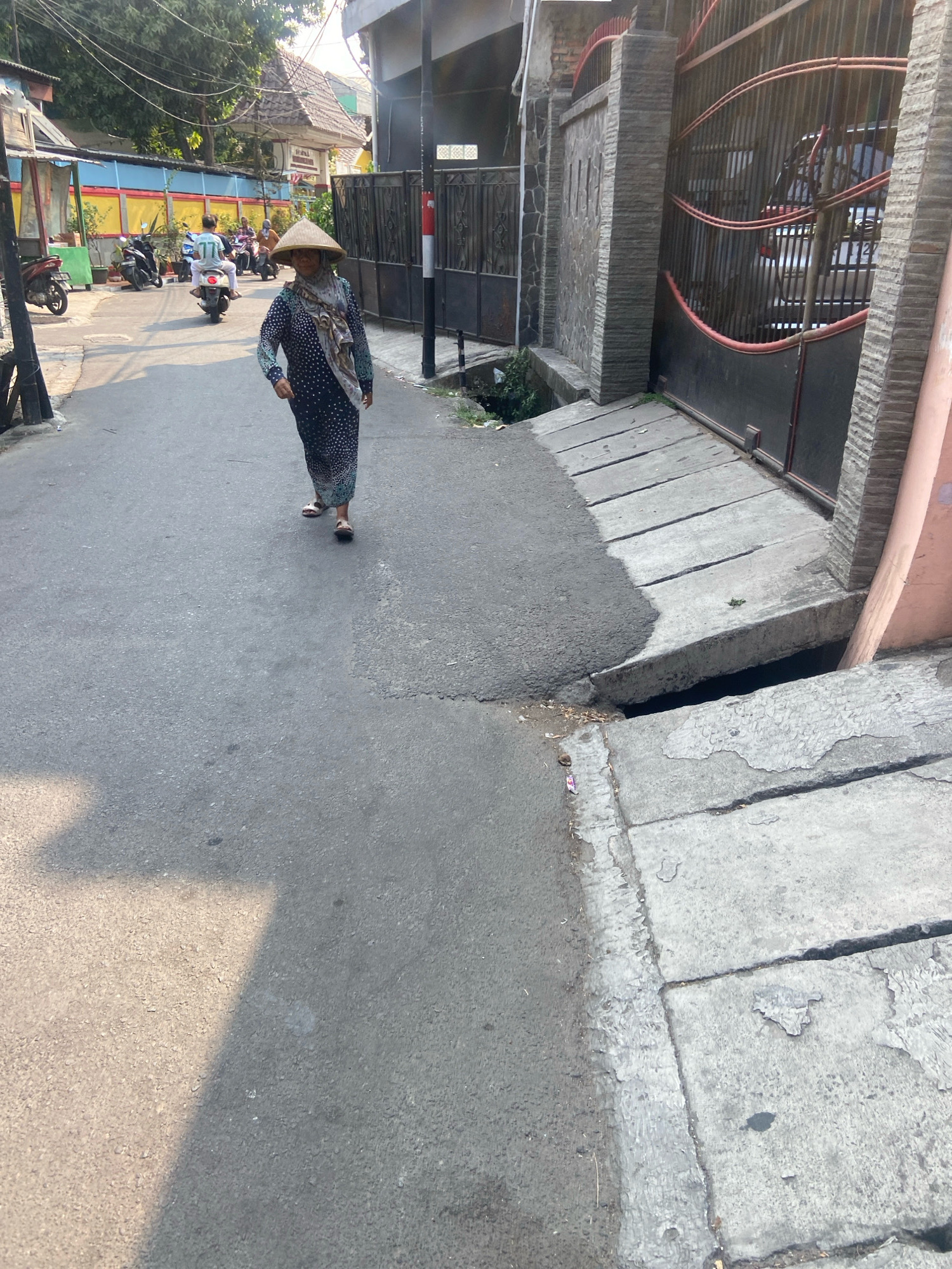 A woman wearing a conical hat and a patterned dress walks down a narrow, paved street in North Jakarta, Indonesia. The street is lined with buildings, one of which has a brightly colored facade. Several motorcycles are parked along the side of the street, and two are in motion further down the road, carrying riders. A closed metal gate with red accents is visible on the right side of the frame, leading to a property. The ground on the right side of the street is a raised concrete walkway with visible cracks and an open drainage channel. The lighting suggests it is daytime, with shadows cast by the sun.
