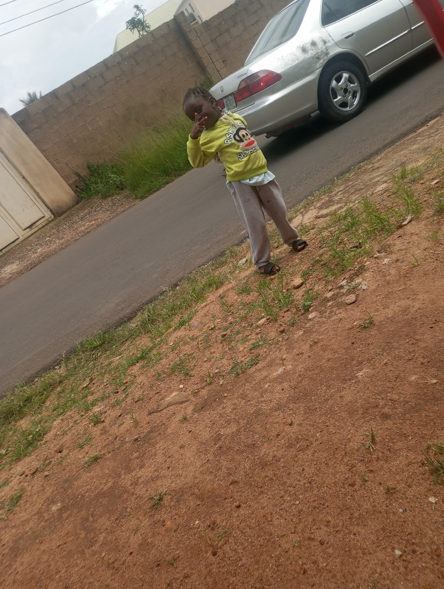 A child, possibly a girl, is standing on a dirt and grass verge next to a paved road. The child is wearing a lime green long-sleeved shirt with a graphic of a monkey and the words "RISKY BUSINESS" and grey sweatpants. The child is making a peace sign with their right hand. Behind the child, a silver sedan car is parked on the road. The car appears to be a Honda Accord. A brick wall runs along the far side of the road, with a beige metal gate visible to the left. The sky is overcast. The scene appears to be located in Jos, Nigeria, given the characteristic red earth and vegetation.