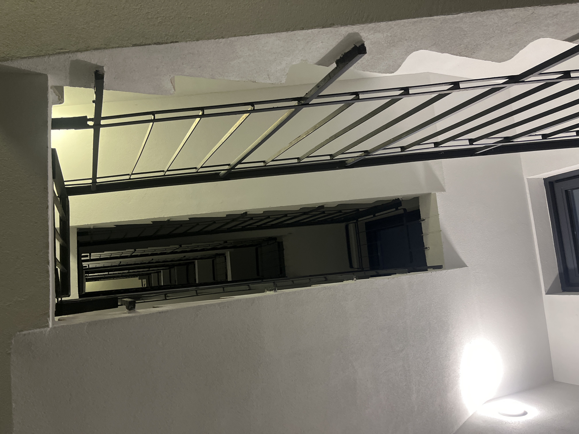 This is an upward shot in a stairwell. The perspective is from a low angle looking up through several stories. There is a metal railing above. The walls are painted white. There is a window on the right side of the frame. The lighting is dim, and there is a circular light fixture on the wall in the lower right. The location is Saint Petersburg, Russia.
