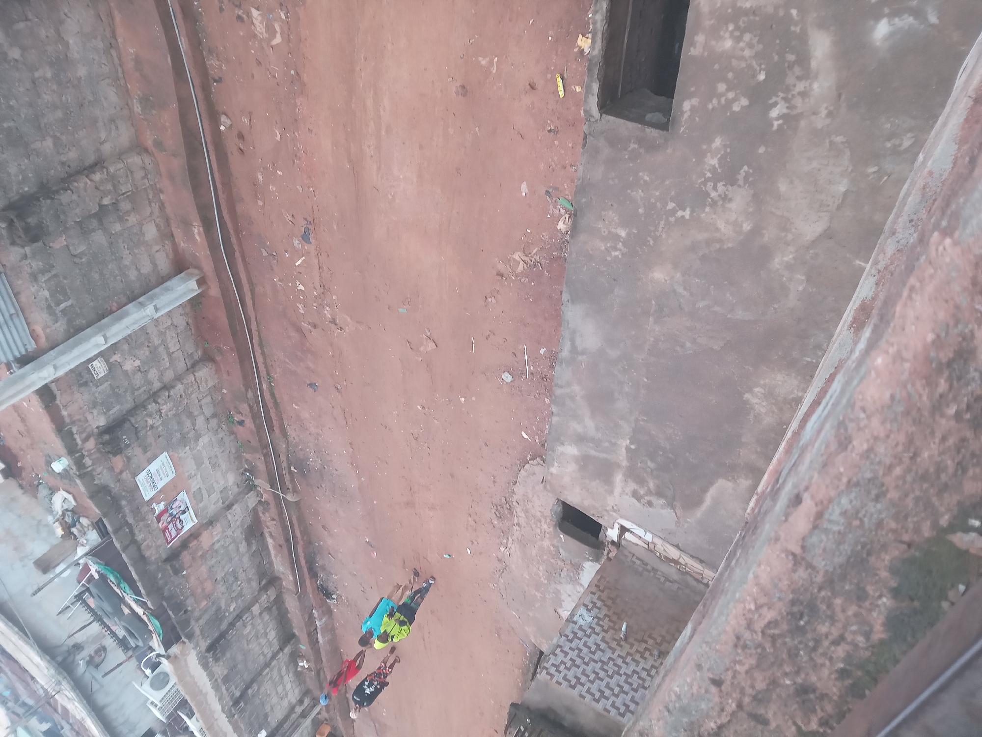 Viewed from above, the image shows a group of four people walking on a dirt path between two buildings in Onitsha, Nigeria. The building to the left is a multi-story structure, possibly residential, with signs posted on the exterior. The building on the right has a weathered, gray exterior. The path is largely dirt with scattered debris.
