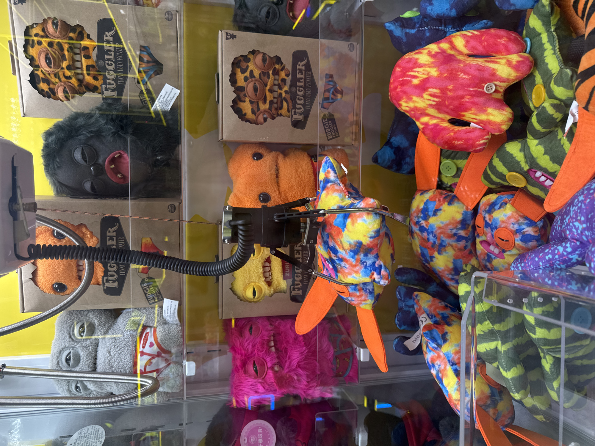 The image shows the inside of a claw machine filled with "Fuggler" plush toys. The toys have varied appearances, including different colors, facial features, and some are in boxes. The claw of the machine is currently holding a blue, yellow, and orange Fugglers toy. Some toys have buttons for eyes or other features. Text on the boxes reads "Fuggler Funny Ugly Monster" and "Budgie Fugglers Edition". A product certification sticker with Chinese writing is visible. The exact location cannot be determined from the image. The image appears to have been taken at an arcade or amusement venue.