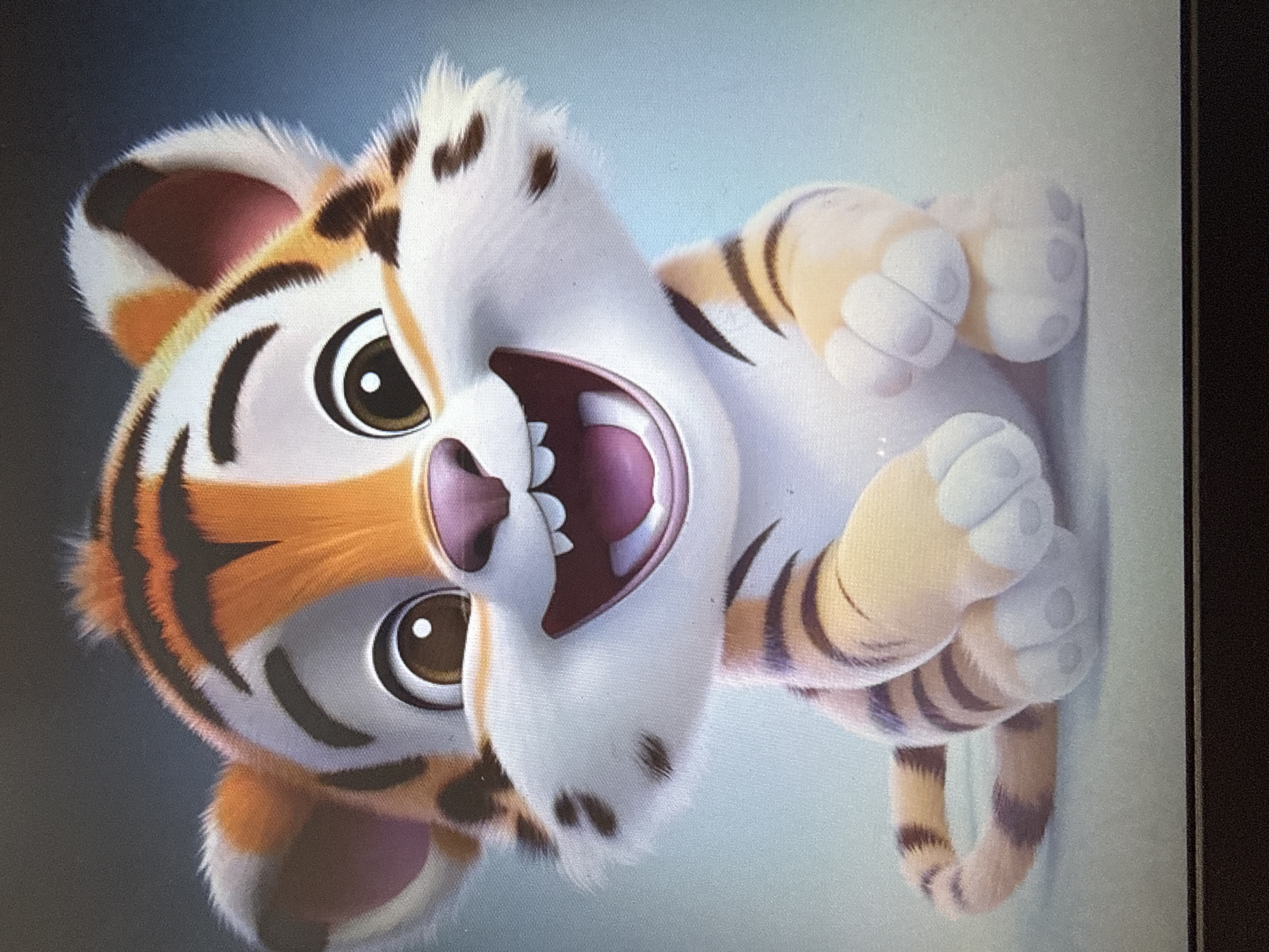 A digital illustration depicts a cartoon tiger cub sitting on a flat, light-colored surface against a softly blurred blue and gray background. The tiger cub is facing forward with its mouth open in a wide, joyful grin, revealing white teeth and a pink interior. It has large, expressive brown eyes with white pupils, a pink nose, and orange and black stripes on its head and body. Its ears are fluffy and white with pink interiors. The cub's body is plump and covered in soft-looking fur, with orange and black stripes on its legs and tail. Its paws are white with grey toe pads. The tiger cub's tail is curled around its body. The image is cropped at the top and bottom, with the bottom edge of a dark, flat object visible at the very bottom of the frame. The scene does not depict any specific city or location within a city.