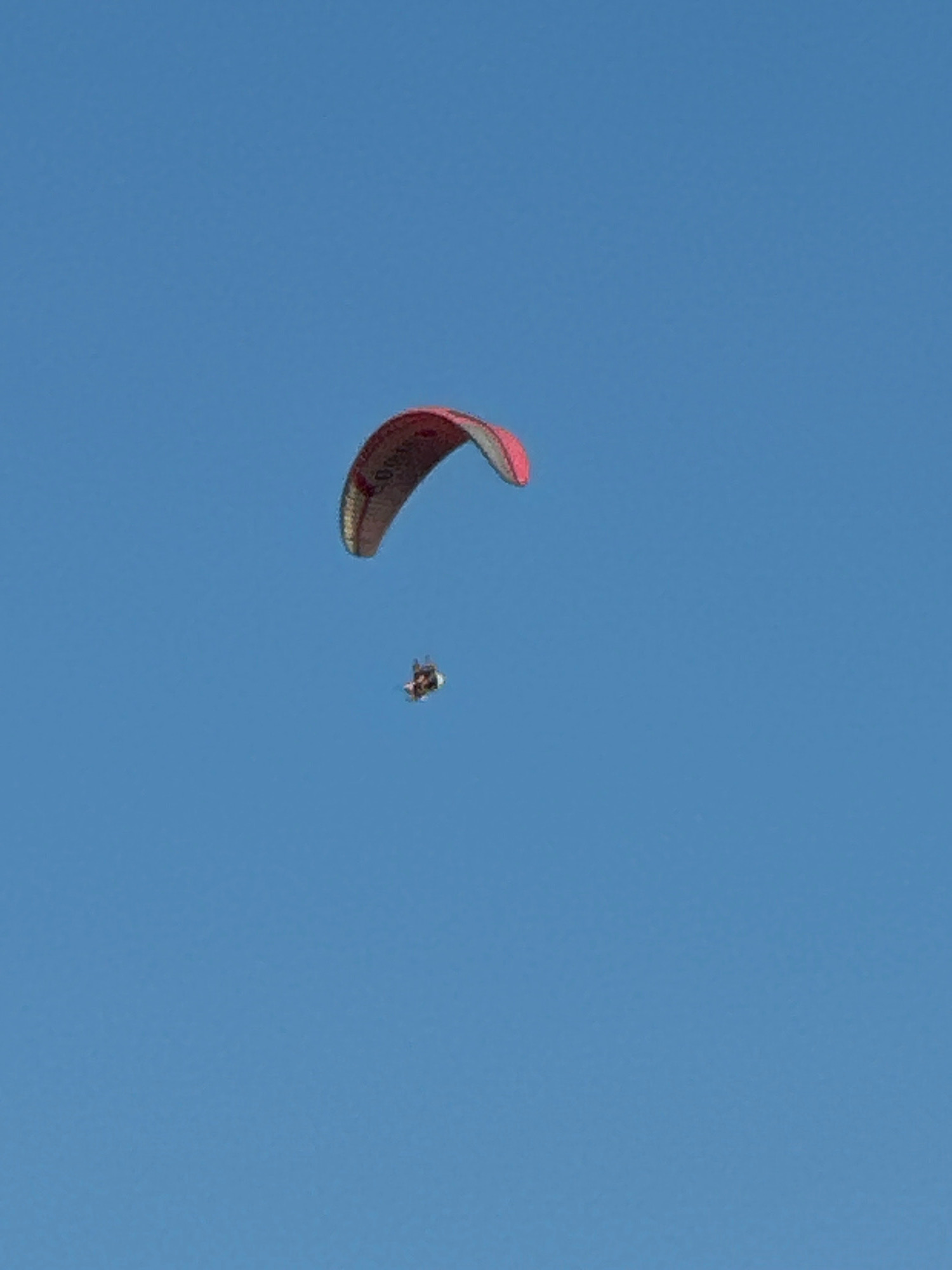 A paraglider with a red and grey wing is in flight against a clear blue sky. Below the paraglider, a much smaller object, likely a second paraglider or a drone, is visible. The image captures a moment of aerial activity in what is presumed to be the sky above Boreti, Montenegro.