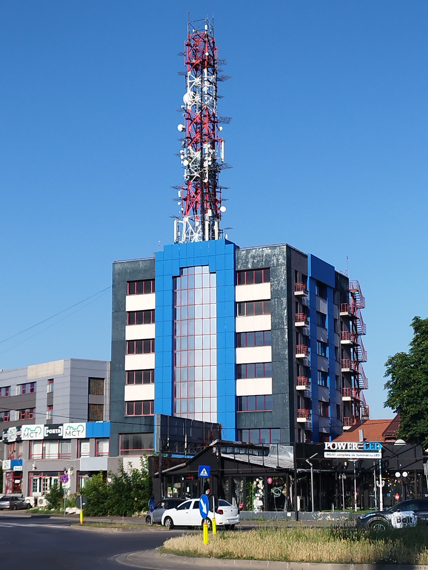 The image shows a tall telecommunications tower attached to a modern, multi-story building with a blue facade. The building is flanked by other structures, including a lower gray building with signs that read "MCT" and "PORTA MCT". In the foreground, there is a street with cars, a sidewalk, and grassy areas. A few people are visible near the sidewalk, and the sky is clear and blue. This scene is located in Bucharest, Romania. Specifically, the building complex with the tower is situated in the northern part of Bucharest, near the intersection of Pipera Boulevard and Fabrica de Glucoză Street. The telecommunications tower is a prominent landmark in this area, serving communication needs for the city. The commercial establishments at the base of the main building, such as "POWER-LED" which sells lighting solutions, indicate a mixed-use urban environment.