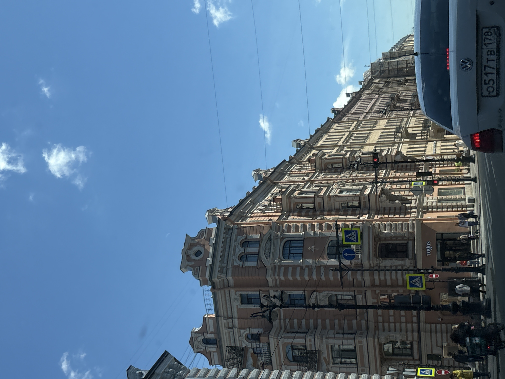 The image shows a street scene in Saint Petersburg, Russia, on a sunny day with scattered clouds. The architecture is characteristic of the city, with ornate buildings lining a wide street. On the left, a multi-story building with a pink facade and decorative white trim features multiple windows and balconies. A lamppost stands prominently in front of it. Further down the street, a row of older buildings with yellow and beige facades continues, displaying intricate architectural details. Pedestrian crossings and traffic signals are visible on the sidewalk. Several people are seen walking along the sidewalk, and a few are near the shops. A white Volkswagen sedan is in the foreground, partially obscuring the view of the street ahead. The license plate is visible and appears to be Russian. The overall impression is that of a bustling urban environment with historical architecture.