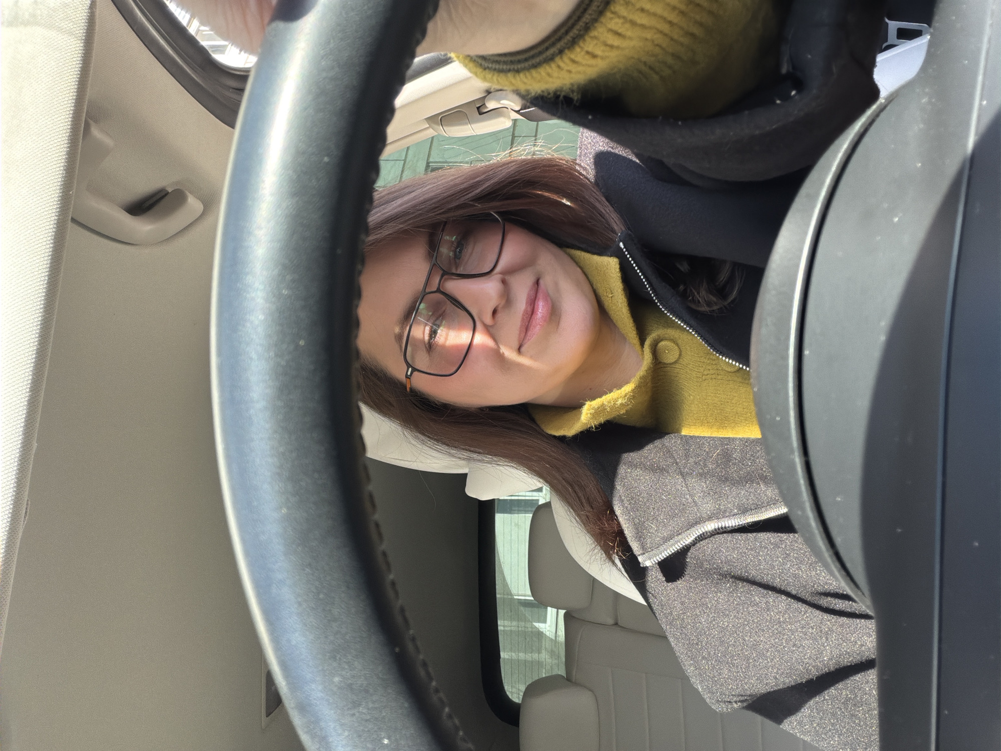 A woman wearing glasses and a mustard yellow sweater with a black jacket is visible from the chest up, seated in the driver's seat of a car. The steering wheel is prominent in the foreground, partially obstructing the view. The car's interior is light-colored. The background visible through the car windows shows a building exterior with windows. The exact location in Chaikovsky, Russia cannot be determined from the image provided.