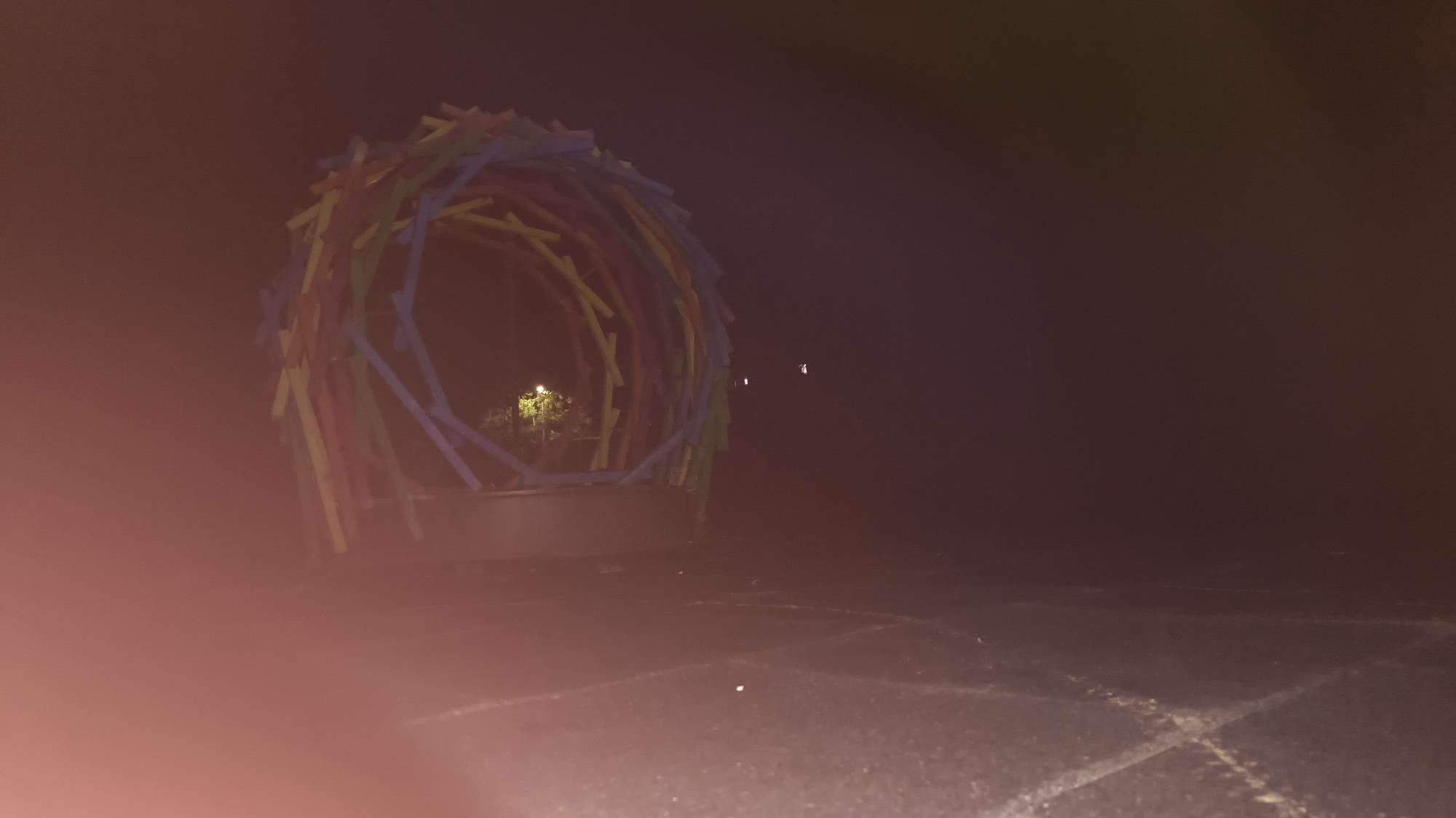 A low-angle, night-time photograph captures a circular, multi-colored structure constructed from what appear to be painted wooden or plastic rods. The structure, positioned on the left side of the frame, is illuminated from within by a warm light, highlighting a dense cluster of green foliage. The ground in front of the structure is a dark, paved surface, possibly asphalt, with faint white markings. To the right of the structure, the background is dark and hazy, with a few small, indistinct lights visible in the distance. A soft, reddish-pink light flares prominently in the lower left corner of the image, indicating a light source outside the frame. The overall atmosphere is dim and slightly mysterious. The location is identified as Mikhaylovka, Russia.