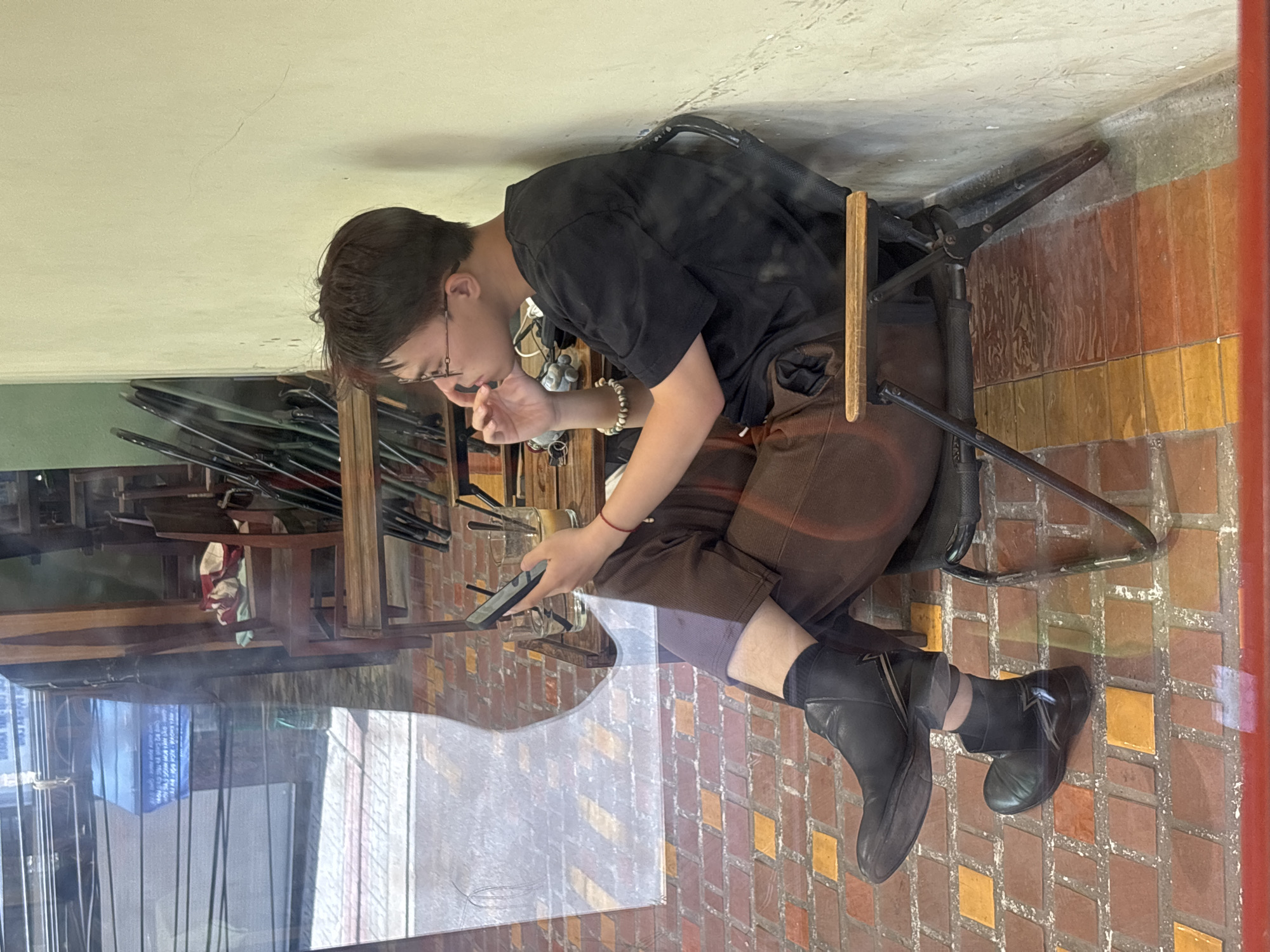 A young man with short, dark hair and glasses sits in a black folding chair against a light yellow wall, looking at his phone. He is wearing a black t-shirt, brown pants, and black boots. A small wooden table with drinks and straws sits beside him. The floor is tiled with red, orange, and yellow squares. The scene appears to be viewed through a window, with reflections showing a street and buildings in Nha Trang Ward, Vietnam.