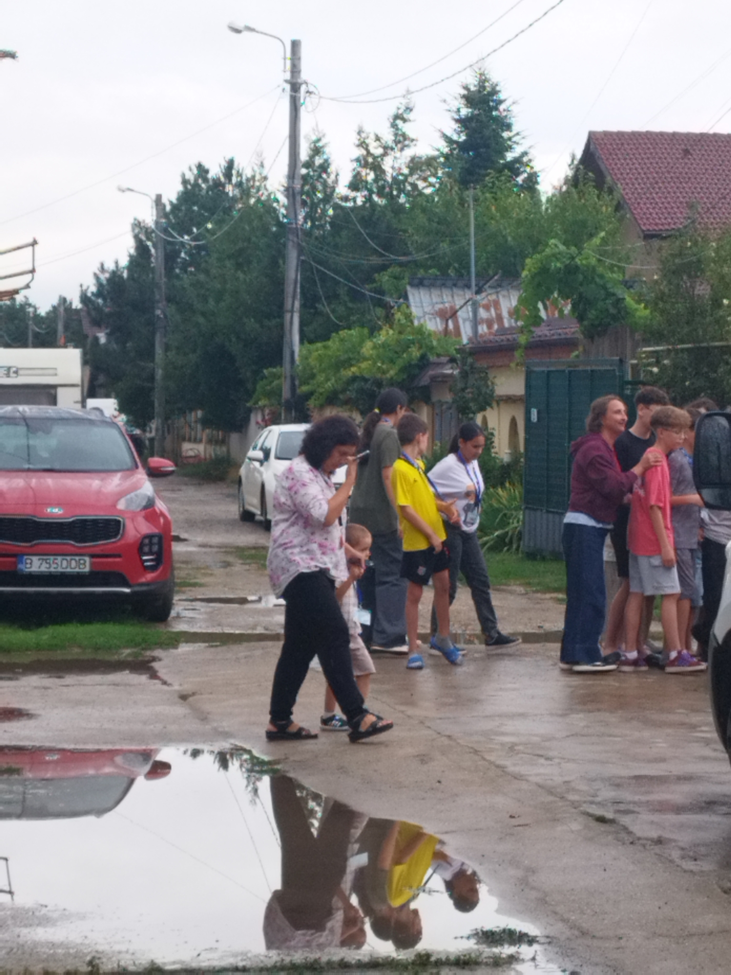The image depicts an outdoor scene, possibly in Bucharest, Romania, on a cloudy day. In the foreground, a red Kia Sportage car is parked on the left side, displaying license plate "B 75500B". A puddle of water reflects the car and the surrounding environment. To the right of the car, a woman in a floral shirt and black pants is walking and holding a phone to her ear, accompanied by a young boy.
Further down the street, a group of people, predominantly children and young adults, are gathered and interacting. Several of the children are wearing bright yellow shirts, while others are dressed in casual clothing. One woman in a maroon hoodie appears to be interacting with a boy in a pink shirt. The background reveals residential buildings with red-tiled roofs, trees, and utility poles with electrical wires. The overall environment suggests a suburban or residential street setting, with wet pavement indicating recent rain.