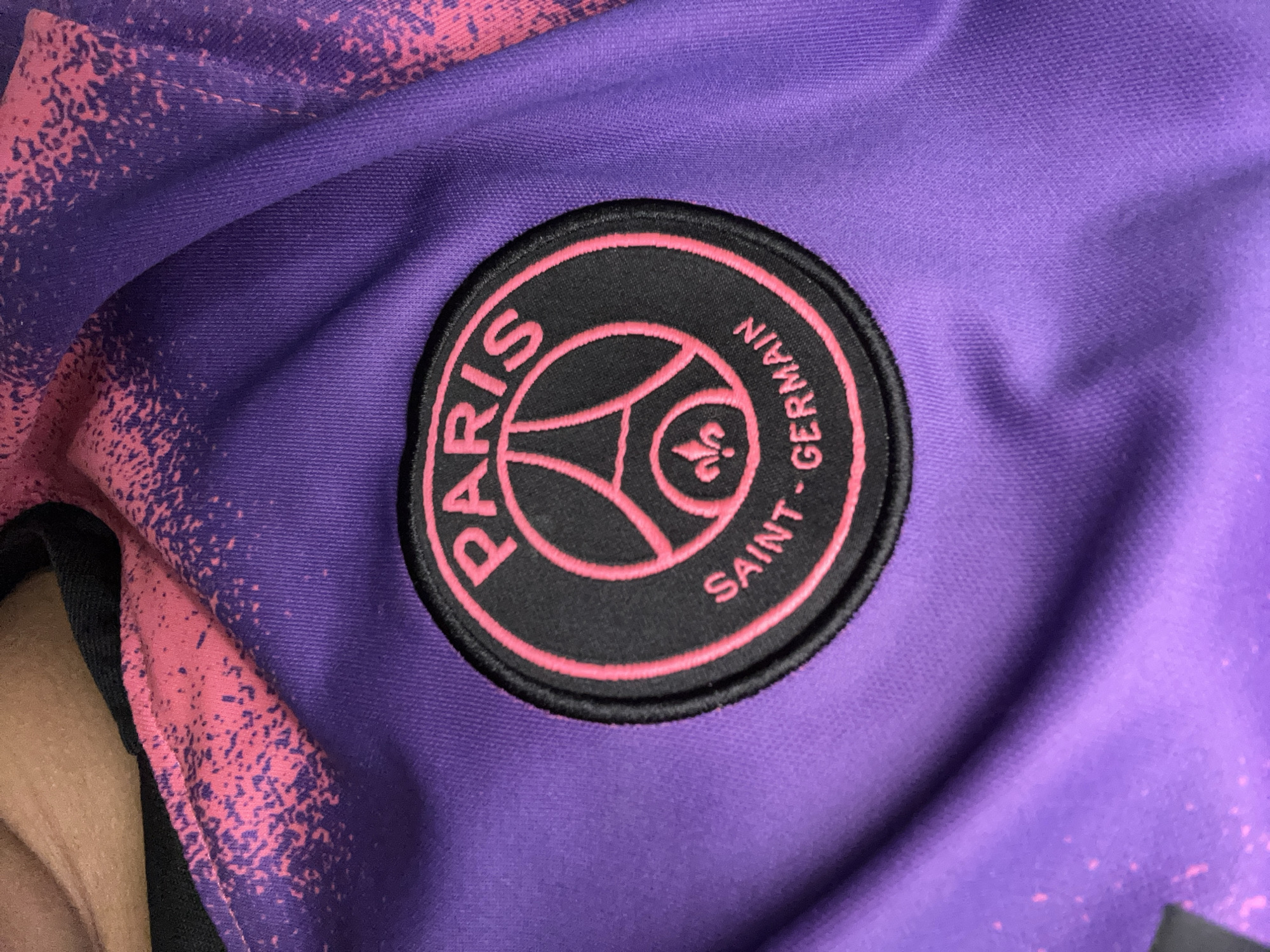The image shows a close-up of a purple fabric, likely a piece of clothing, with a round black patch featuring the Paris Saint-Germain logo. The logo is outlined in pink and contains the Eiffel Tower and a fleur-de-lis. The words "PARIS" and "SAINT-GERMAIN" are written in pink around the emblem. The fabric has a speckled pink and purple pattern in the upper portion of the image, and some of the fabric is creased, suggesting it is being worn or is folded. The background is a blurred expanse of the same fabric. There is no information about the city or location in the image.