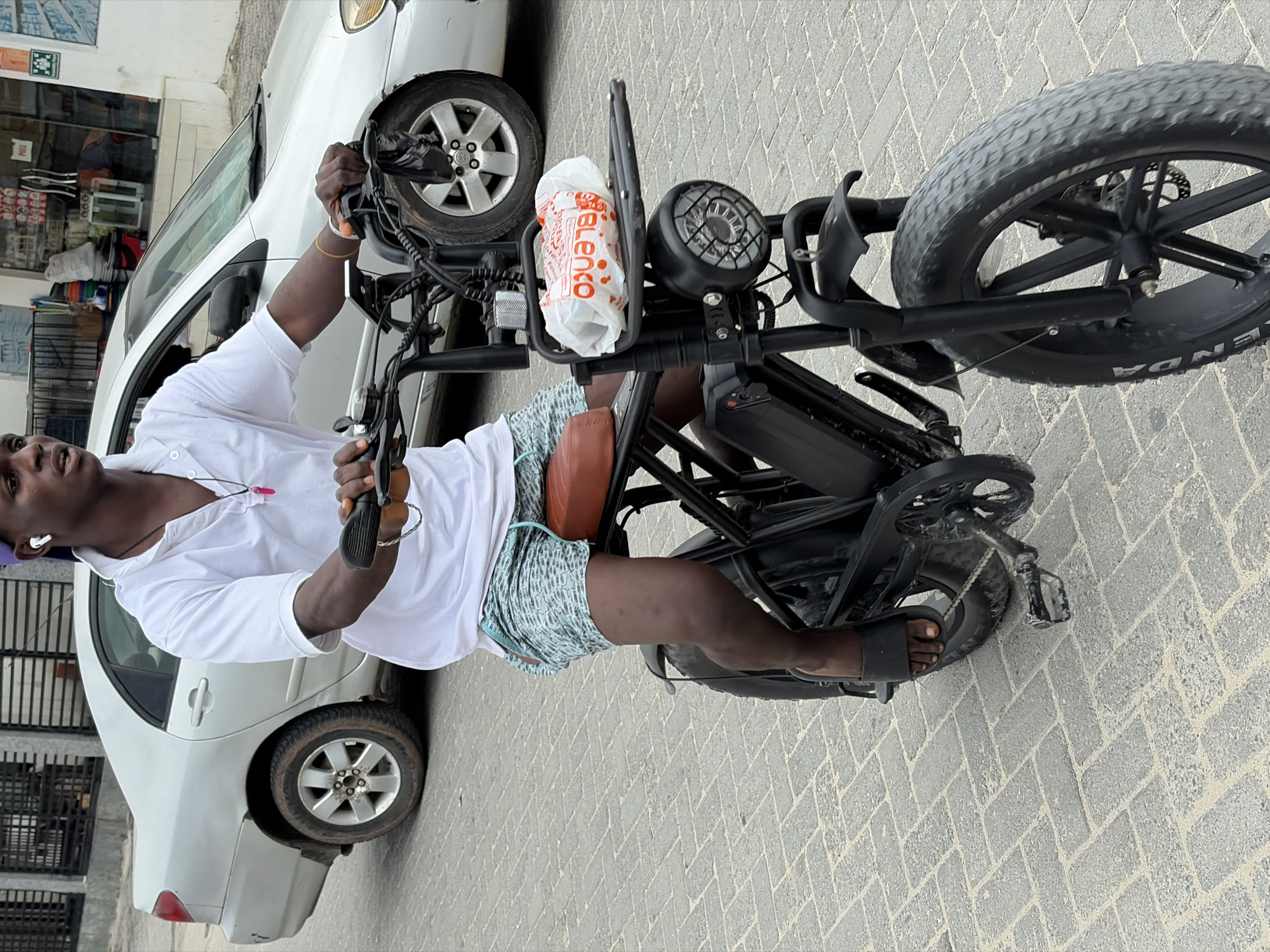 A man is seated on a black electric bicycle in Igboefon, Nigeria. He wears a white button-down shirt, patterned shorts, and black sandals. The bicycle has a large front headlight and a bag from "Blenko" secured to the front. A silver car is parked next to the bicycle. The ground is paved with herringbone pattern bricks.