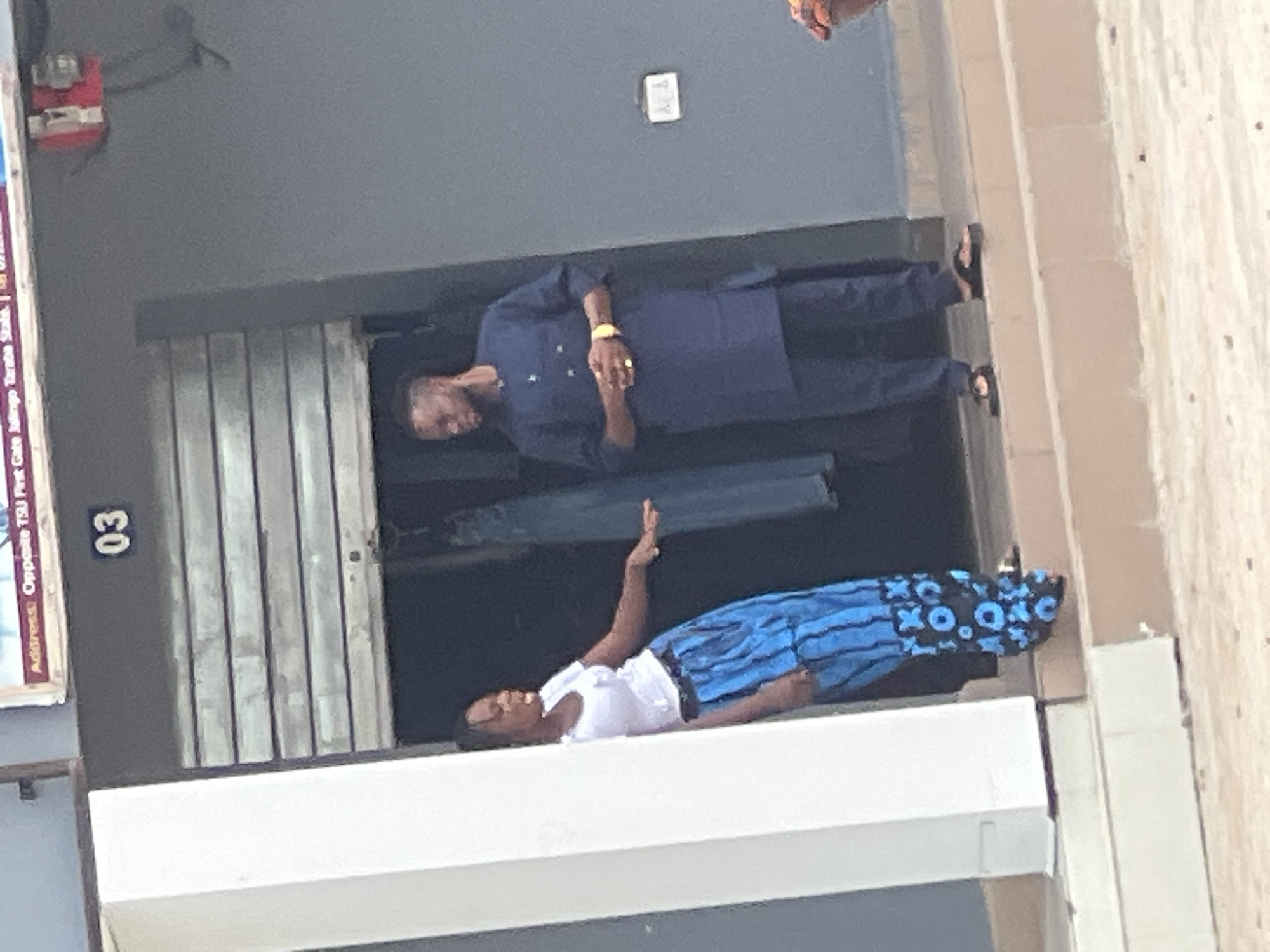 The image depicts a storefront in Jalingo, Nigeria. A woman in a white t-shirt and a blue patterned skirt is standing on the left, gesturing with her right hand. On the right, a man in a dark blue traditional outfit stands with his hands clasped. A metal roll-up door is visible behind them, with the number "03" displayed prominently above it. The storefront appears to be located on a street with a sidewalk and steps leading up to the entrance. A red electrical box is mounted on the wall to the right of the door. The overall scene suggests an urban commercial setting.