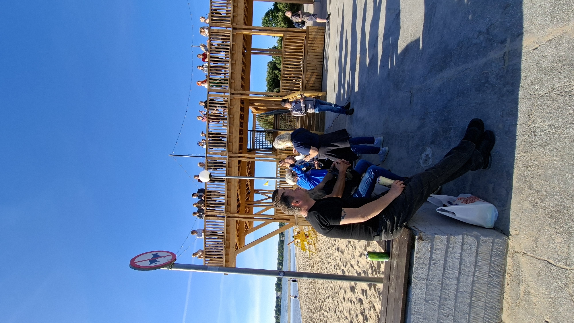 The image captures a sunny day scene in Tallinn, Estonia, with a clear blue sky dominating the upper portion. In the foreground, a man in a black t-shirt and dark jeans sits on a wooden bench, facing left, with a green can placed to his left. Next to him, two other individuals are seated. Behind them, a group of people stand on a multi-level wooden structure.
The wooden structure appears to be a viewing platform or a cafe with seating areas, featuring railings and stairs. Several people are visible on the different levels of this structure, some appearing to be standing and conversing, while others are leaning over the railings. A sign with a red circle and a diagonal line over a silhouette of a dog is mounted on a pole to the left of the wooden structure, indicating a "no dogs allowed" rule.
The background reveals a sandy beach area with beach chairs. Further in the distance, a body of water, likely the Baltic Sea, is visible, with a hazy shoreline on the opposite side. The ground in the foreground is a paved area with visible shadows cast by the people and the structure, suggesting the sun is positioned to the right and slightly behind the viewer. A white shopping bag rests on the ground beneath the bench. The overall impression is a relaxed, outdoor gathering at a coastal location.