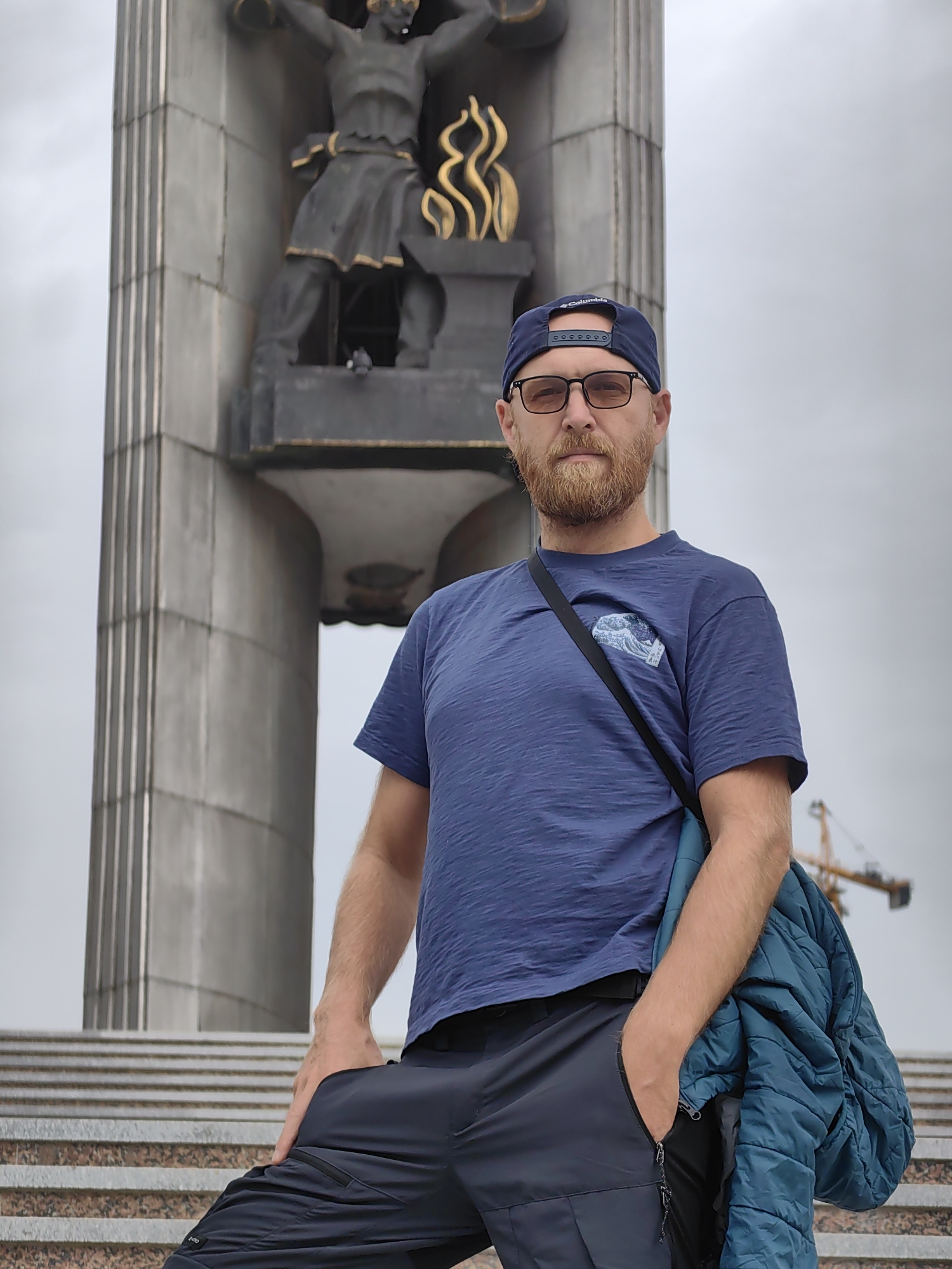 A man stands on steps in front of a large monument, likely in Izhevsk, Russia. He wears a blue t-shirt, black pants, a blue baseball cap, and glasses. He has a red beard. A jacket hangs over his shoulder, and one hand is in his pocket. The monument behind him features a statue of a man holding a structure with golden flames. A crane is visible in the background. The sky is overcast.