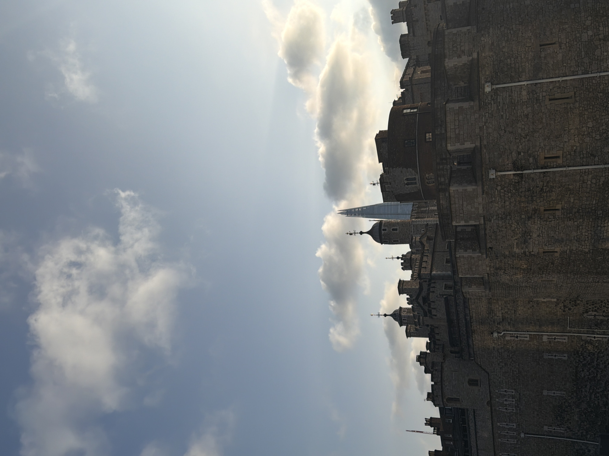 The image shows the Tower of London in London, United Kingdom. It's a stone castle complex with multiple towers, including the White Tower, a central and iconic structure. Some portions appear very detailed, while other areas are blurred. The shard skyscraper is partially visible. The sky is light blue with fluffy clouds.