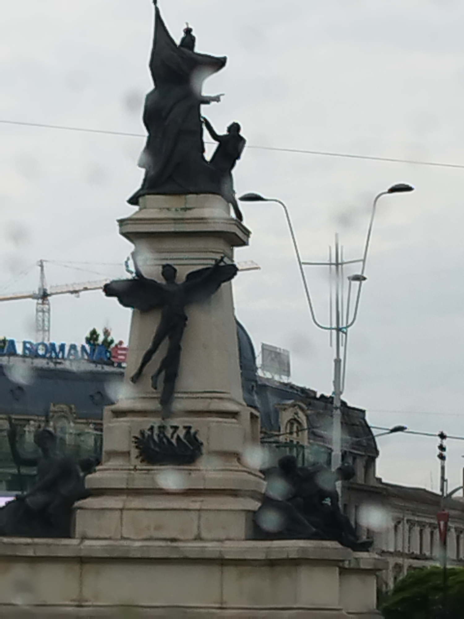 The image shows a monument in Bucharest, Romania, under a cloudy sky. The monument features a large central statue of a figure holding a flag and pointing, with smaller statues surrounding its base. One of these smaller figures appears to be an angel with wings, reaching upwards. Another figure is depicted as a fallen soldier. The monument is made of stone and bronze. In the background, buildings with architectural details are visible, along with a construction crane and streetlights. The date "1877" is inscribed on a wreath on the monument. The overall scene suggests a historical or commemorative site within the city.