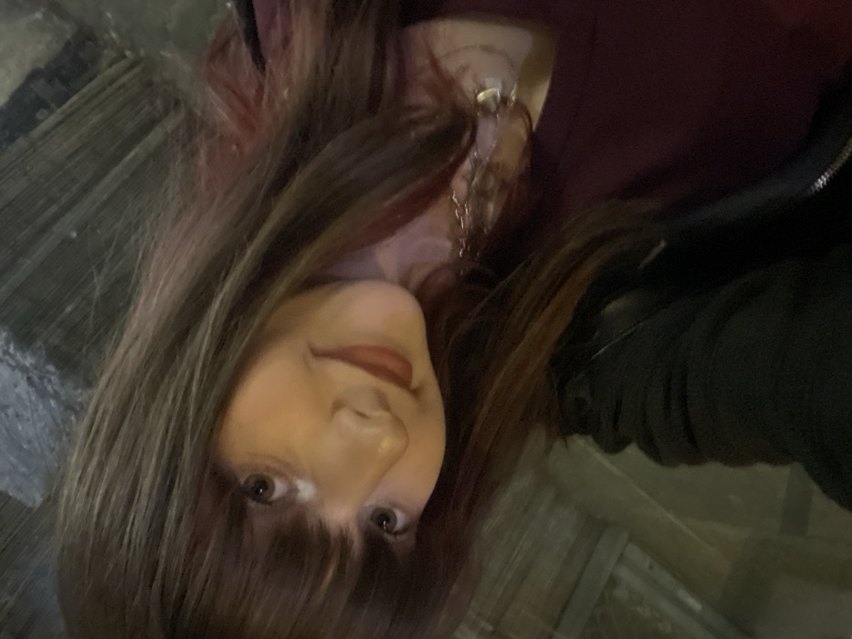 A person with long brown hair and bangs is facing the camera. They are wearing a maroon shirt and a black jacket. A silver necklace is visible. The background appears to be outdoors, possibly on a street, with some visible textured surfaces and dark areas. The image is slightly tilted.