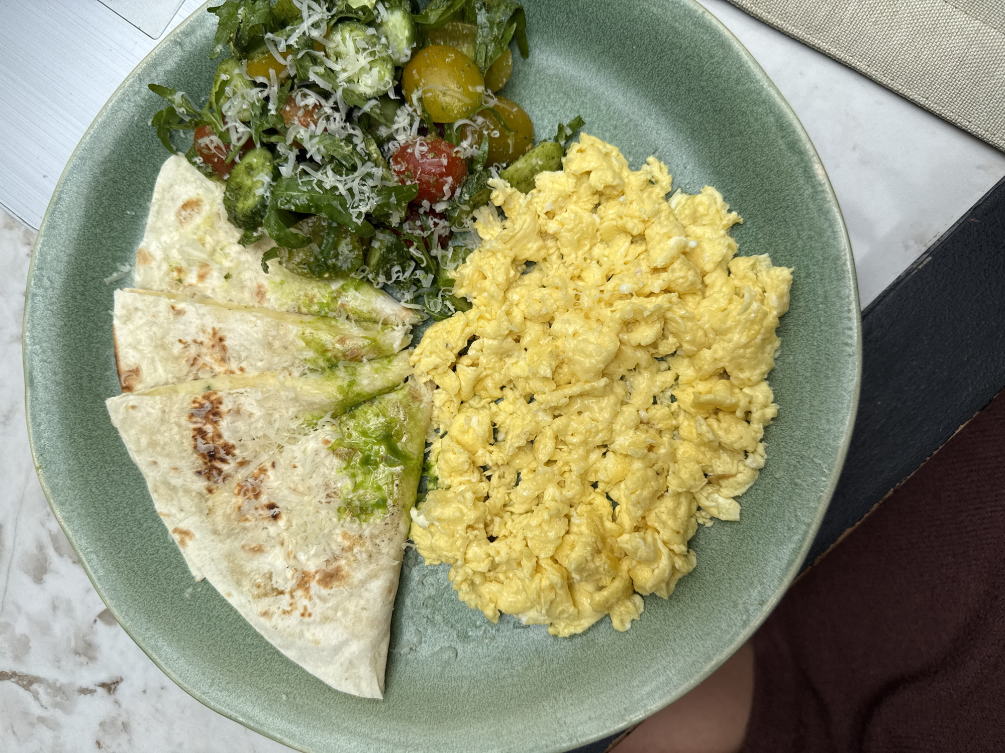 A green plate containing scrambled eggs, sliced quesadillas, and a salad with tomatoes and shredded cheese rests on a surface. The quesadillas are topped with a green sauce, possibly pesto. The salad includes green and red tomatoes, along with other greens. The surface appears to be a table with multiple textures and colors, including white and gray marble, a beige material, and a brown covering. The location is not discernible from the image.