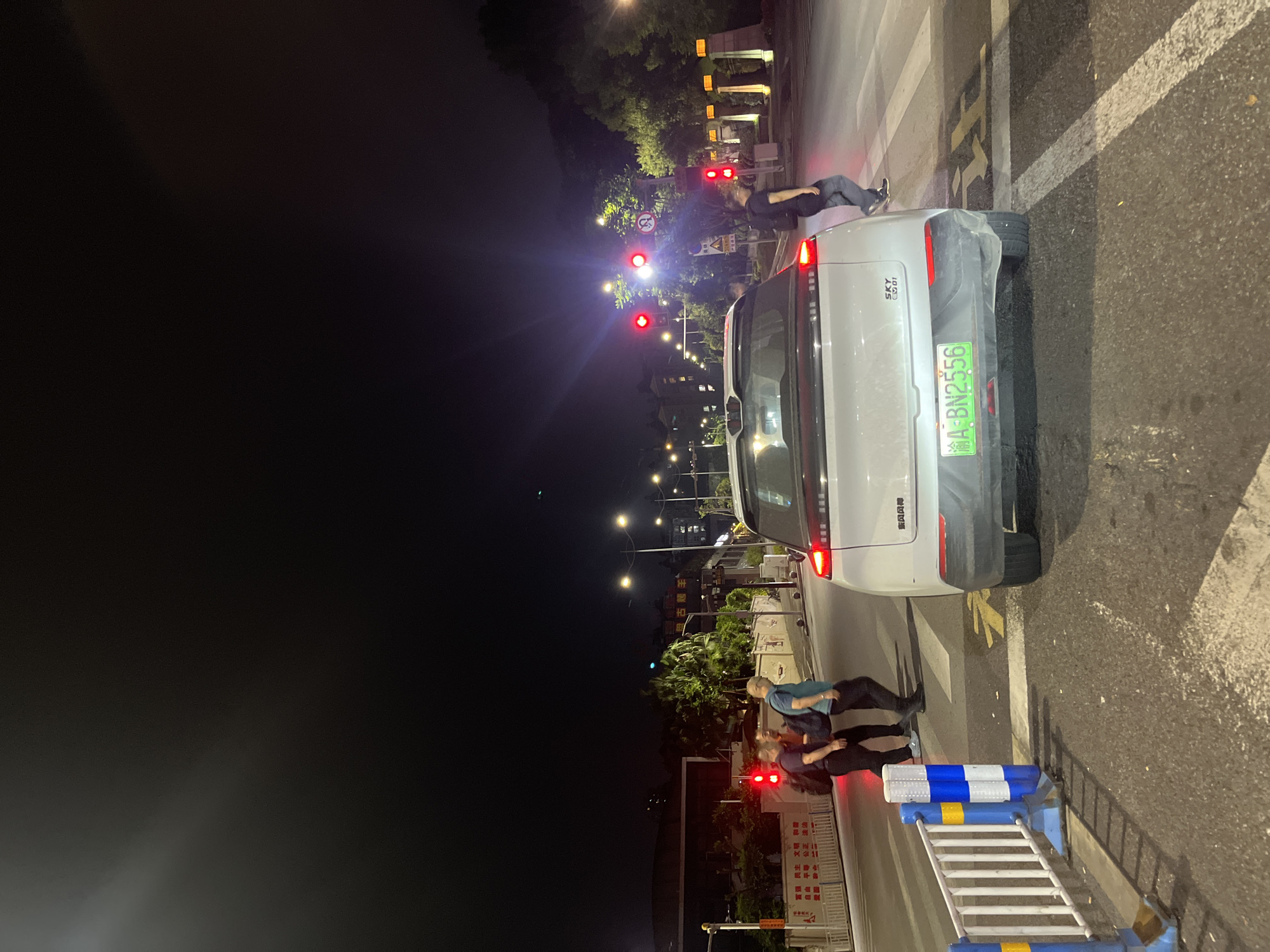 At nighttime on a street in Jiulongpo District, China, a white SUV is stopped at a pedestrian crossing. Three pedestrians are visible on the left side of the road, crossing the street. One person, wearing a black shirt and dark pants, is walking with a black backpack. Beside them, another person, wearing a dark blue shirt and dark pants, also carries a backpack. The third person, in a blue shirt and dark pants, walks slightly ahead of the others. Traffic lights on the left show red, indicating a stop. On the right side of the road, another pedestrian, dressed in a black shirt and dark pants, is crossing the street. The SUV has a license plate that reads "A-BN2556". Streetlights illuminate the scene.