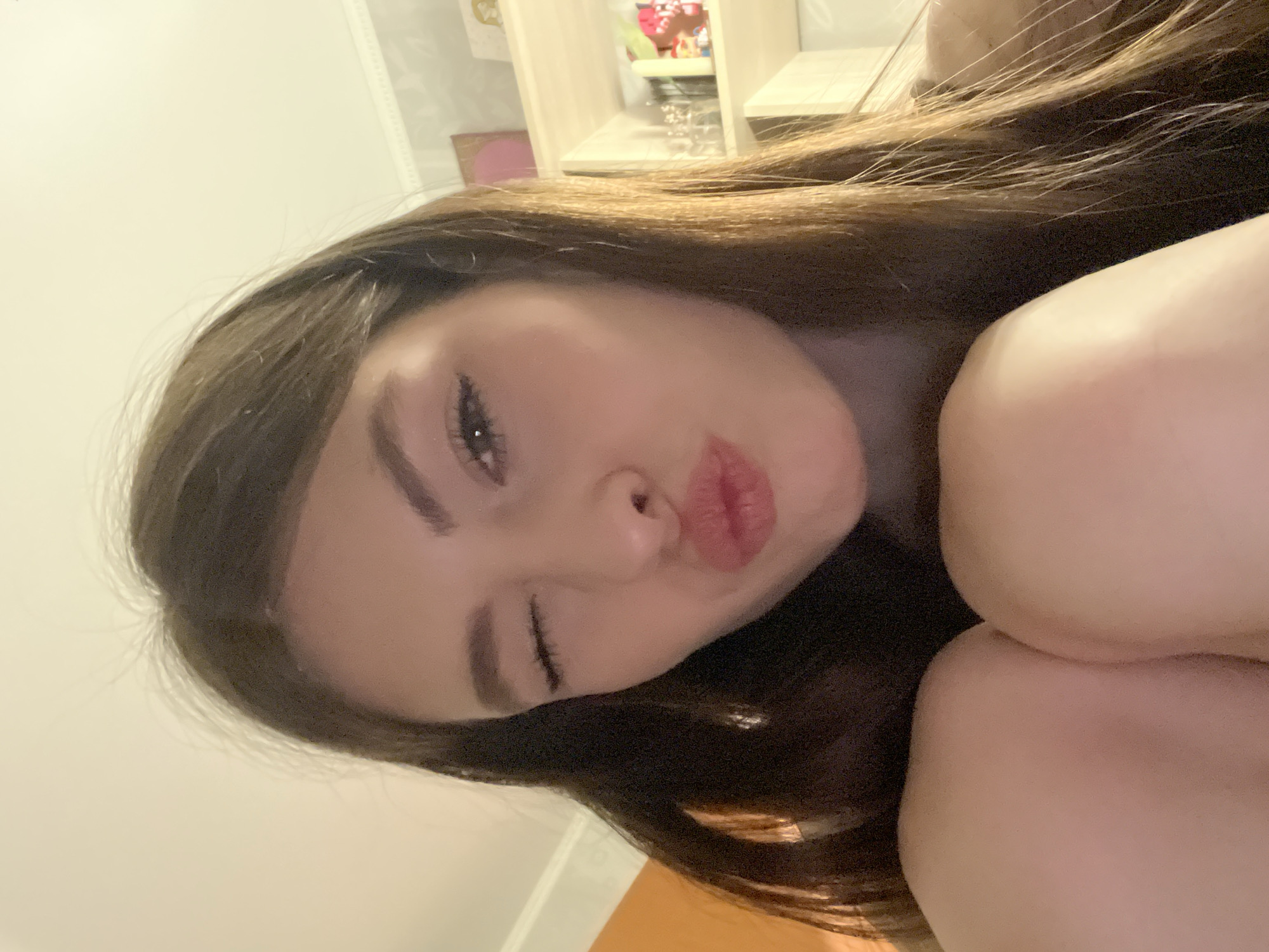 A close-up, horizontally oriented portrait features a young woman with long, dark brown hair. She is positioned casually, possibly lying down or leaning, with her face dominating the left side of the frame. Her facial expression is playful and affectionate: she is winking with her right eye (left from the viewer's perspective) and puckering her lips in a kissy face. Her eyebrows are well-defined, and she has visible eyeliner and mascara.
Her dark brown hair flows around her head, with some lighter brown or auburn highlights visible, particularly near the top of her head. The image is framed tightly, showing parts of her upper arms or legs, which are light-skinned, creating soft, rounded forms in the foreground and right side of the image.
The background is softly blurred but reveals an indoor setting. On the left, a light-colored wall is visible. Towards the upper right, there appears to be a light wooden shelf or cabinet with a few indistinct items, suggesting a personal room or living space. The lighting is soft and warm, hinting at an indoor setting, possibly during the evening or with artificial light. No text is visible in the image. The overall mood is intimate and cheerful.