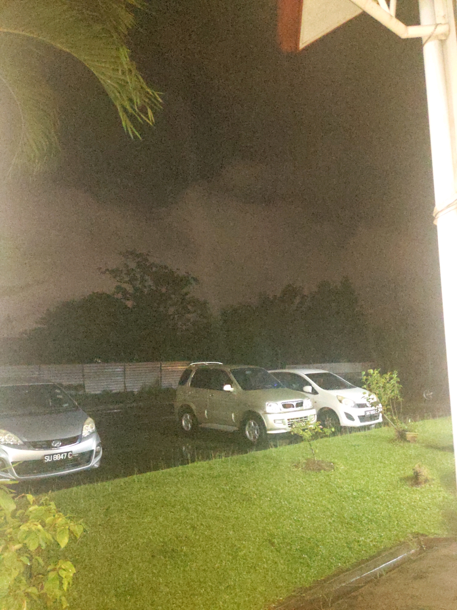 The image depicts an outdoor scene at night, possibly during rain, in Kolombong, Malaysia. In the foreground, a well-maintained green lawn is visible, with a dark drainage channel running across it. Two cars are parked side-by-side on a paved surface in the mid-ground. The car on the left is silver and has the license plate "SU 8847 C". The car on the right is white and its license plate is partially obscured but appears to read "SAC 9853 C". Behind the cars, a metal fence runs horizontally, and beyond that, dense foliage and trees are visible, silhouetted against a dark, cloudy sky. A large palm frond overhangs the scene from the top left. A structure with white columns and an angled roof is present on the far right of the image. The overall lighting suggests it is nighttime, with artificial lights illuminating the vehicles and the white structure. The atmosphere appears hazy, possibly due to rain or humidity.
