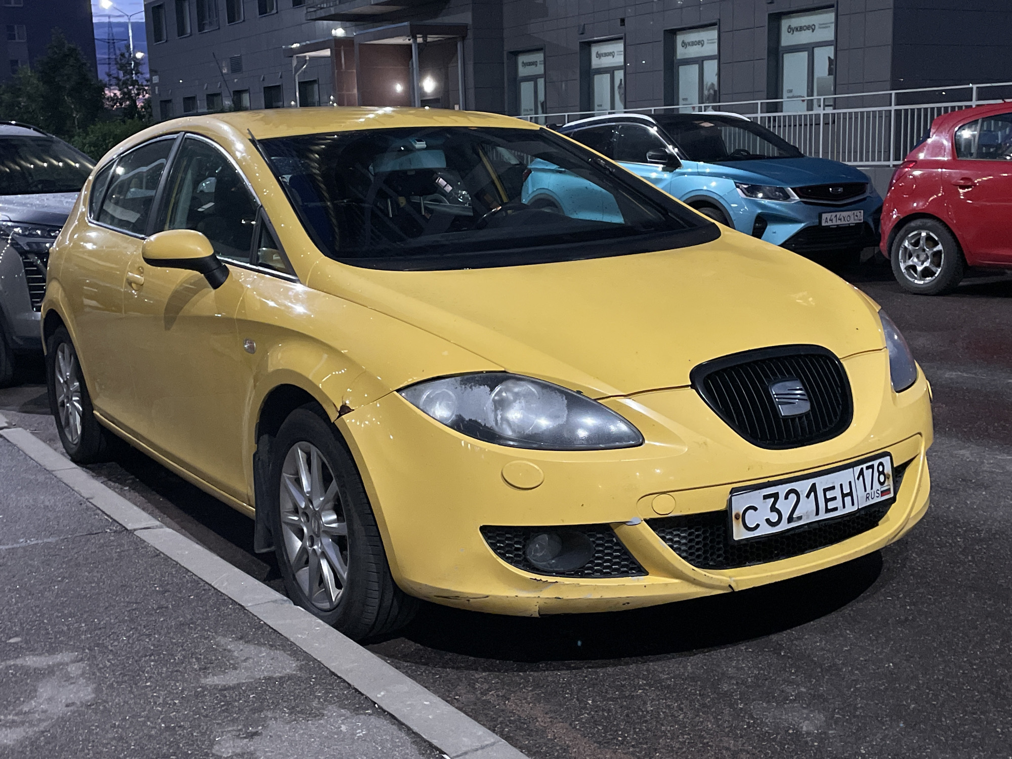 This is a horizontal, medium shot photograph taken at night in Sinyavino, Russia. The main subject is a bright yellow Seat Leon hatchback, parked on a street with a grey curb and asphalt. The car is facing slightly to the right, with its front bumper and headlights prominently displayed. It has a black grille with the Seat logo and a Russian license plate that reads "C321EH 178 RUS". The car appears to have some cosmetic damage on its front fender and bumper.
In the background, several other cars are parked. To the left of the yellow car is a dark-colored SUV. Further in the background, a blue Geely Coolray and a red hatchback are visible, parked in front of a modern building with multiple windows and commercial signage. The windows of the building have signage that appears to be in Cyrillic, with the word "Буквовед" (Bukvoved) visible. The lighting suggests it is nighttime, and the ground appears wet, indicating recent rain or damp conditions. The scene is urban, with the building and parked cars suggesting a residential or commercial area. No people are visible in the frame.