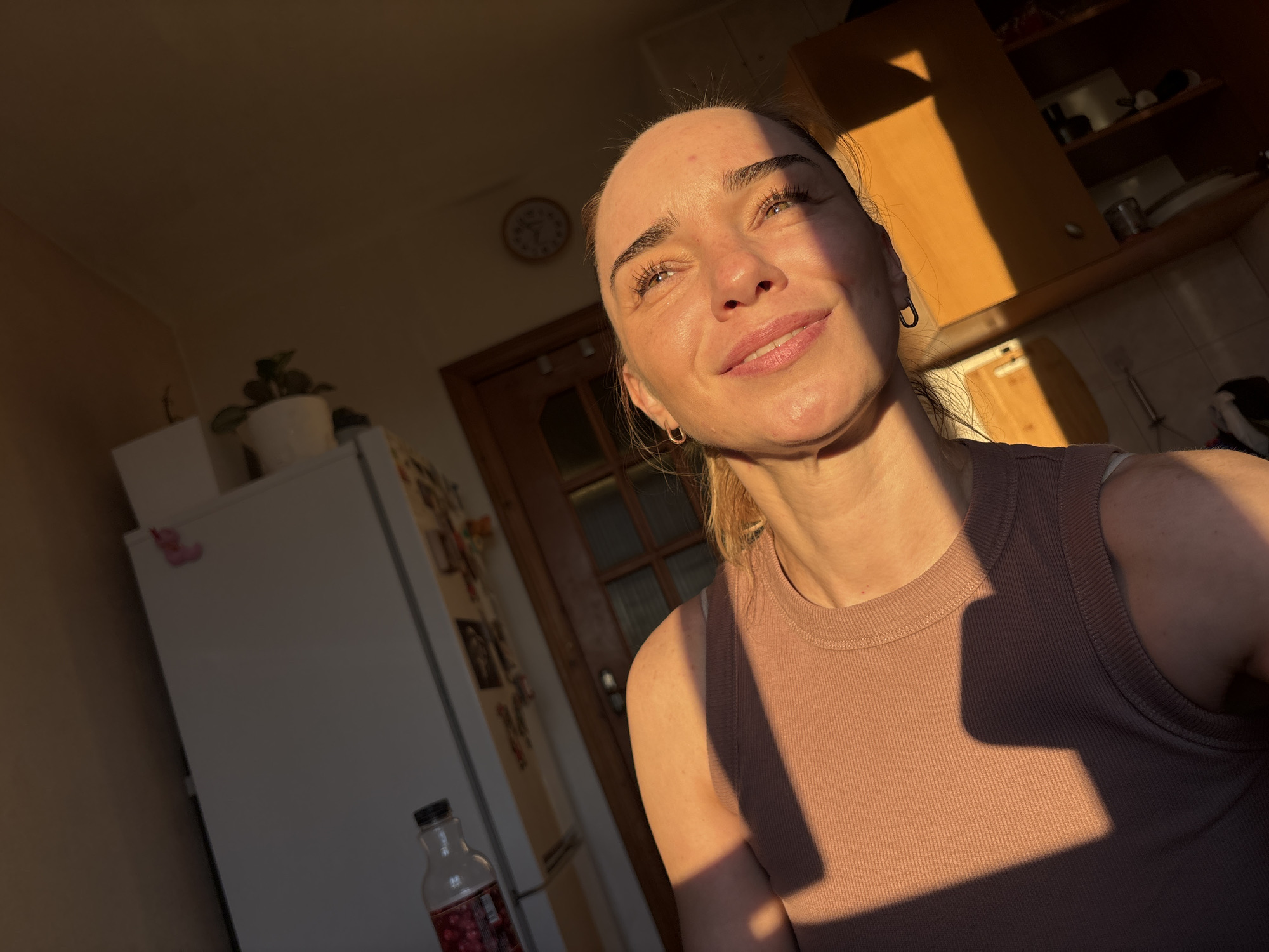 An indoor scene captures a woman in her late 20s or early 30s, with light brown hair pulled back, looking upwards and to the right. She is illuminated by the warm glow of golden hour sunlight, which casts dramatic shadows across her face and shoulders. She wears a simple, ribbed, taupe-colored tank top. Her facial features are accentuated by the light, highlighting her eyes, nose, and lips. A faint smile plays on her lips, and her eyes seem to be looking towards a light source.
In the background, a kitchen environment is partially visible. To the left, a white refrigerator is present, adorned with a pink decoration. A potted plant sits on top of the refrigerator. Further back, a wooden door with glass panels is visible, and beyond that, kitchen cabinets are sparsely stocked with dishes and containers. A clock with a circular face is mounted on the wall above and behind the woman's head. The overall impression is one of a domestic setting bathed in the soft, atmospheric light of late afternoon or early morning. The location is identified as an unknown city in Russia.