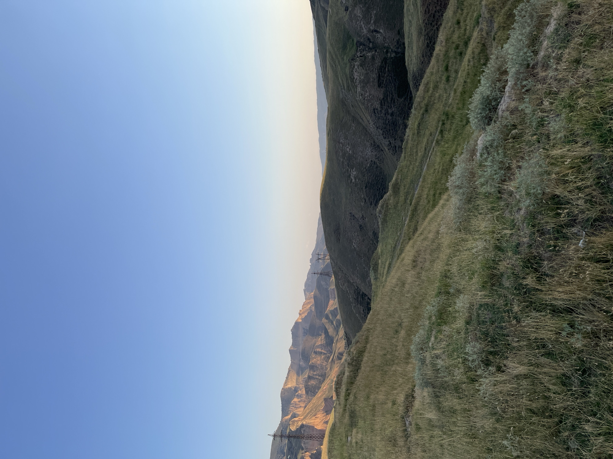 The image shows a landscape in the foothills of Mailuu-Suu City, Kyrgyzstan. The foreground is dominated by dry, golden-green grass and some low-lying shrubs. The middle ground features rolling hills covered in similar vegetation, with a path visible winding up one slope. In the distance, rugged mountains with exposed rock faces are illuminated by the warm, soft light of what appears to be either sunrise or sunset. Two tall, lattice-style electrical towers are visible in the middle ground, suggesting the presence of power infrastructure in the area. The sky is a clear, pale blue, transitioning to a lighter hue near the horizon. There are no people or animals present in the image. The overall impression is one of a remote, natural landscape with man-made elements integrated into it.