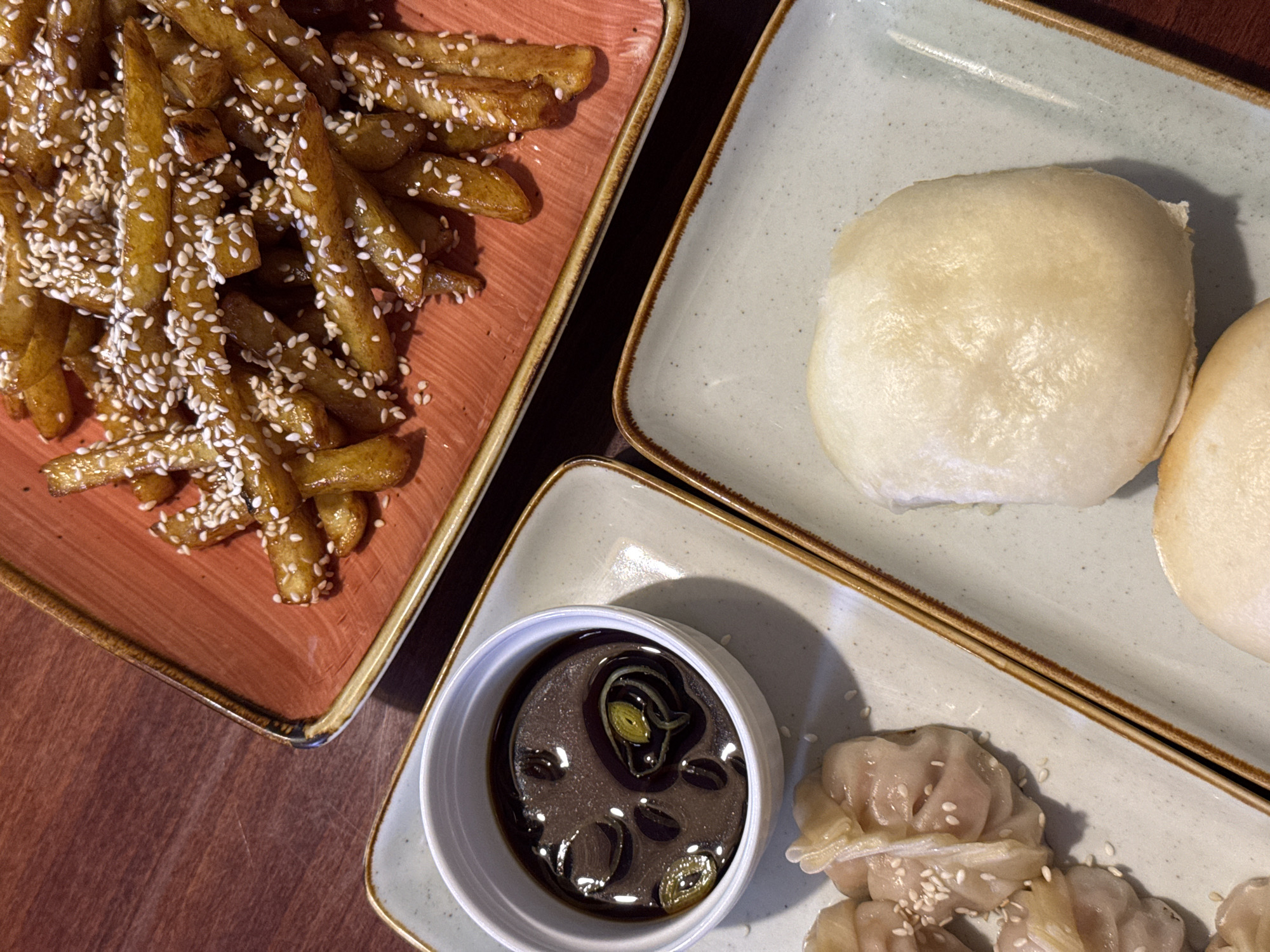 An overhead shot captures a selection of Asian-inspired dishes laid out on a dark wooden table. In the upper left, a reddish-brown rectangular plate holds a generous serving of golden-brown french fries, generously coated in a glossy sauce or glaze and heavily sprinkled with white sesame seeds.
To the right, on a light gray-green rectangular plate with a speckled texture and a brown rim, sit two soft, round steamed buns, often referred to as bao buns. Below these, another identical light gray-green rectangular plate presents several steamed dumplings, each delicately folded and also garnished with white sesame seeds. Accompanying the dumplings is a small, white ceramic bowl filled with a dark dipping sauce, featuring slices of green onion and a subtle sheen on its surface.
The scene depicts a meal being served, likely in an indoor restaurant setting in Podgorica, Montenegro. The overall presentation is neat and appealing. No text is visible in the image.