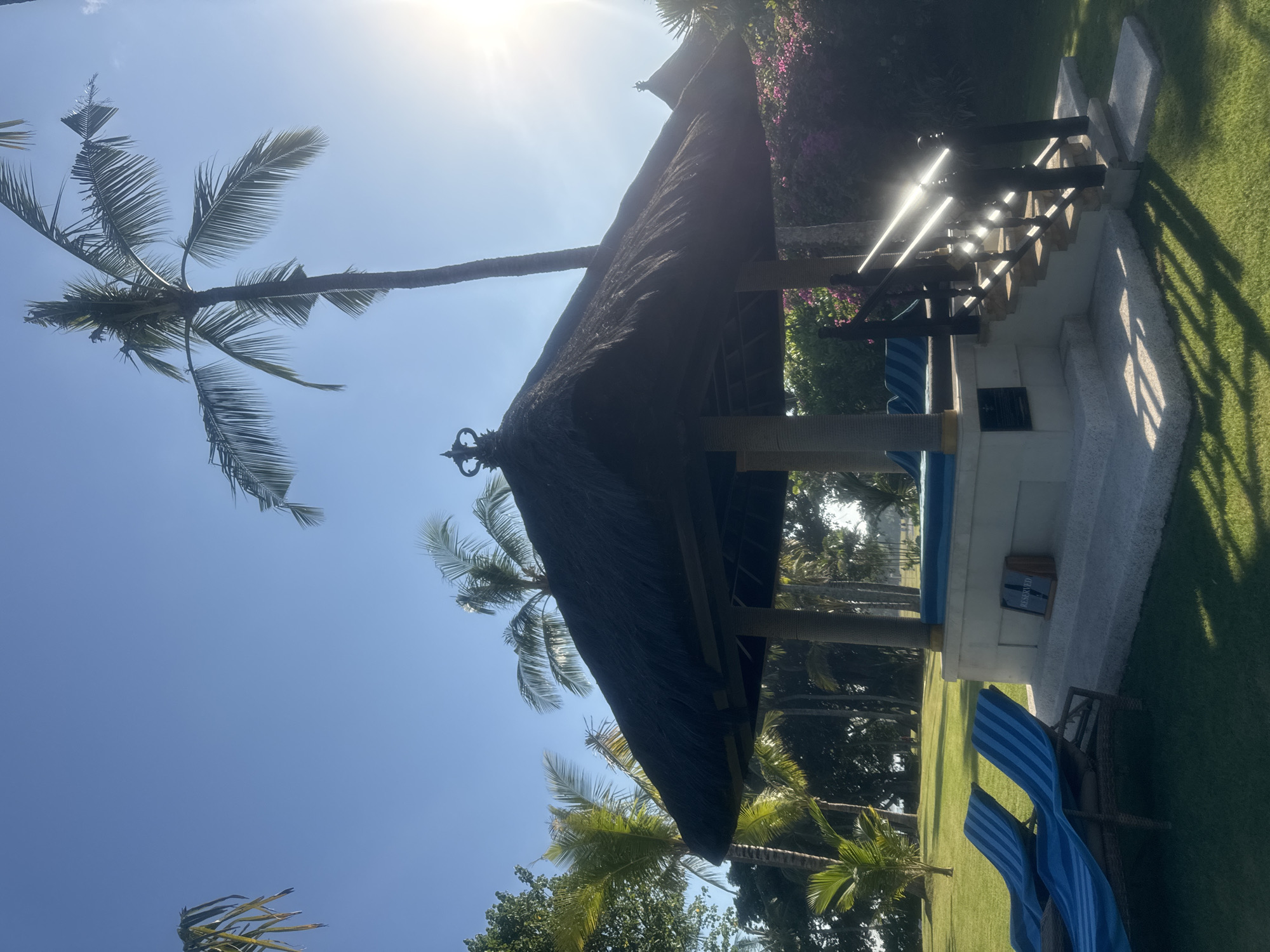 The image captures a bright, sun-drenched tropical outdoor scene, characteristic of a resort or private garden in Kuta Selatan, Indonesia. The main subject is a traditional-style hut or gazebo featuring a dark, densely thatched roof, adorned with an ornate finial at its peak. The hut is supported by dark wooden columns and appears to offer a shaded retreat.
Adjacent to the hut is a small, light-colored concrete structure with steps leading up to an elevated platform. The entire ground area is covered with well-maintained, vibrant green grass, which shows distinct shadows cast by the strong sunlight. Several tall palm trees with broad fronds are visible throughout the frame, some extending into the clear, bright blue sky, while others are part of the dense tropical foliage in the background.
The sun is high and intense, positioned in the upper left, causing a significant lens flare and illuminating the scene with powerful light. This indicates a mid-day setting under clear, sunny weather conditions. Two blue and white striped lounge chairs are partially visible on the grass in the foreground, positioned for relaxation. On the side of the concrete structure, a small sign is visible with the word "RESERVED" clearly printed in white text on a dark background. The scene conveys a sense of tranquility and an inviting tropical environment.