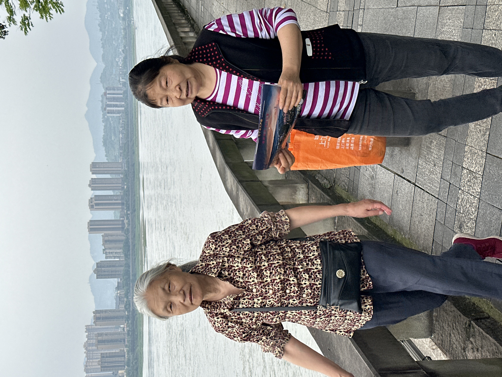 Two mature women stand alongside a river or waterway in the city of Jiangjin, China, on a day with overcast skies. The woman nearest the camera has gray hair and wears a brown and tan patterned blouse, navy pants, a black purse, and maroon shoes. She extends her right arm. The woman next to her has dark hair and is wearing a white and purple striped shirt under a black vest, as well as dark pants. She is holding a brochure or pamphlet and an orange bag printed with Chinese characters. In the background are tall buildings lining the riverbank.