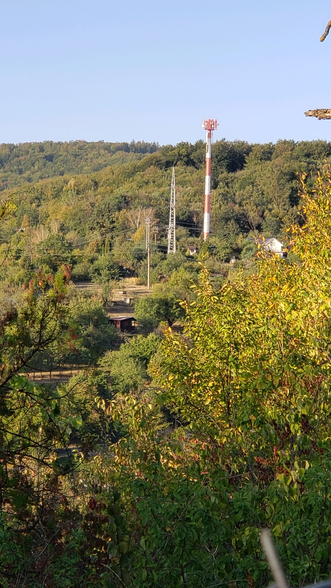 The image depicts a rural or semi-rural landscape with rolling hills covered in dense green trees. A tall, red and white striped communication tower stands prominently in the middle ground, to the right of a shorter, lattice-style tower. Power lines and poles are visible, suggesting infrastructure in the area. Several small structures, possibly sheds or cabins, are scattered among the trees, and a white house is visible further up the hill. In the foreground, a dense canopy of trees with vibrant green and yellowing leaves frames the scene. The sky is clear and blue, indicating fair weather. The image appears to be taken during daylight hours. The location is believed to be in the vicinity of Prešov, Slovakia, likely a wooded area on the outskirts of the city.