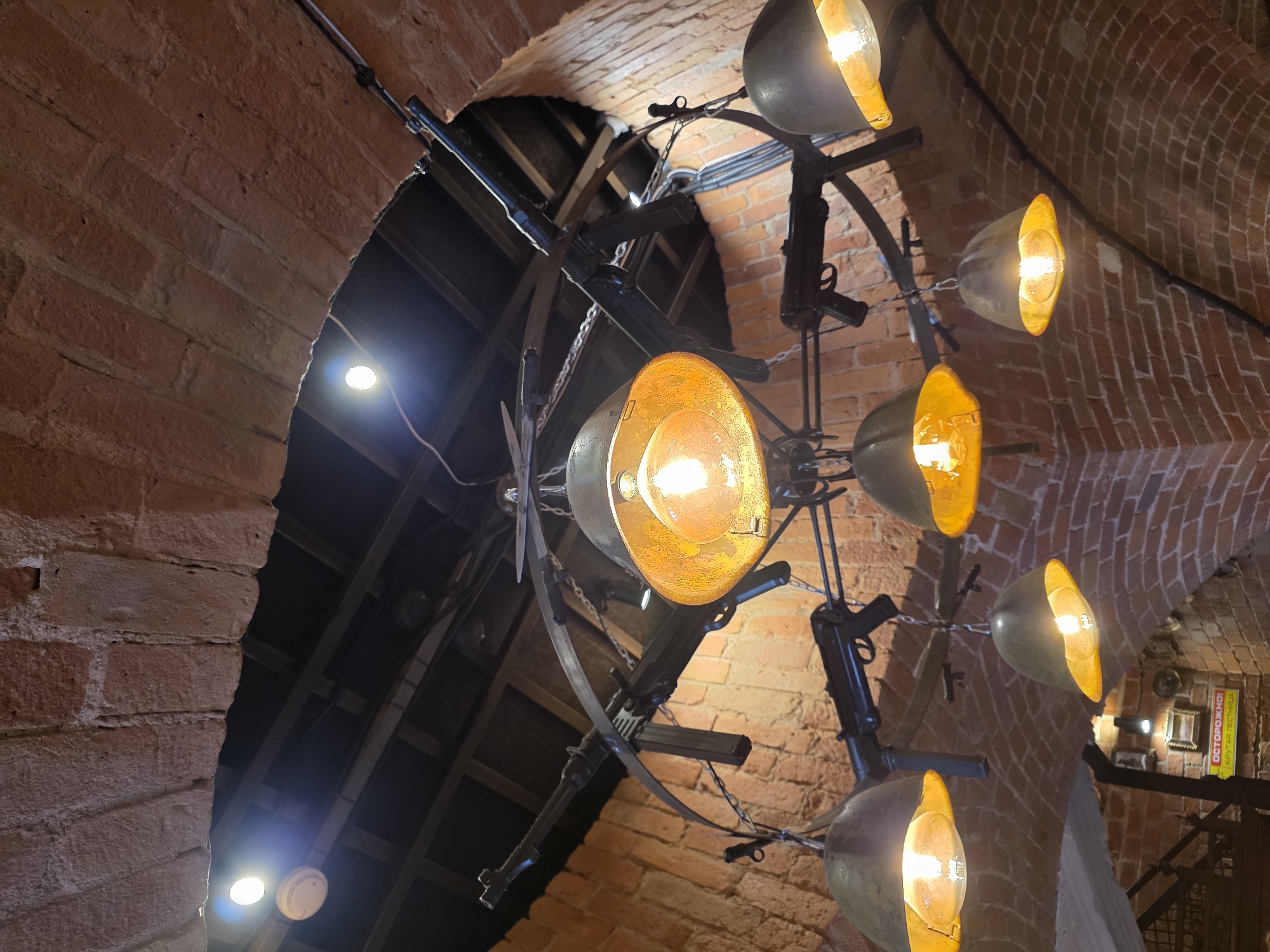 The image shows a chandelier made of what appear to be replica firearms, hanging from a brick ceiling. The chandelier has multiple lamps with warm lighting, and the firearms are arranged in a radial pattern. The setting appears to be an interior space with exposed brick walls. Based on the "ОСТОРОЖНО! КРУТАЯ ГОСТИНИЦА" sign in the lower left of the image, which translates to "CAUTION! COOL HOTEL" and the presence of the guns, this is likely inside the "Kruglaya Bastion" (Round Bastion) museum or restaurant complex in Kaliningrad, Russia, which is known for its historical military exhibits and theme. There are no people visible in the image.