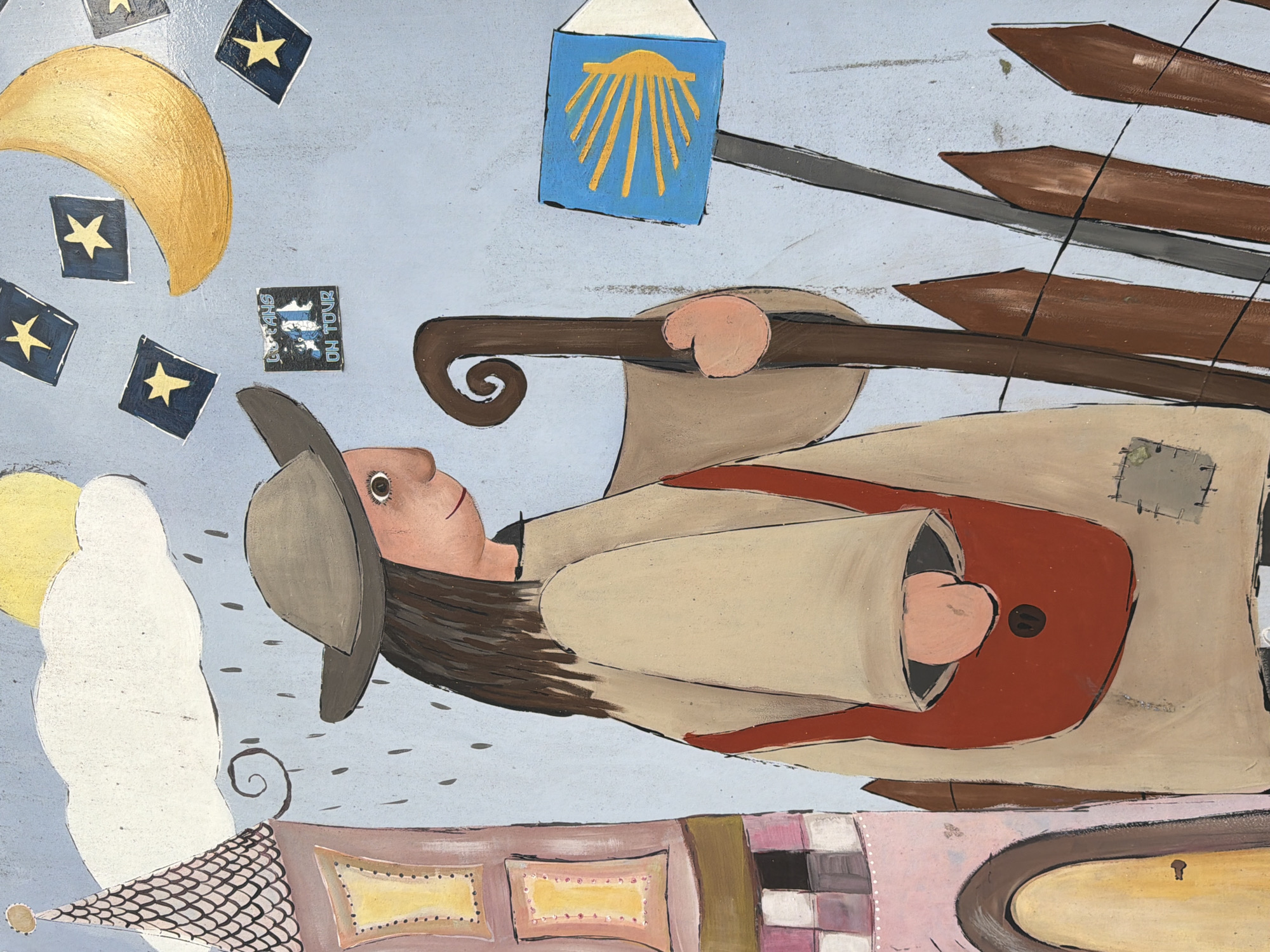 The image is a painting depicting a figure walking in the rain. The figure is dressed in a tan robe, a grey hat, and carries a brown staff with a curled handle. A red bag is slung across the figure's chest. In the background, a yellow sun peeks through a white cloud, and rain streaks are visible. To the left is a pink tower with a conical roof and yellow-framed windows. To the right, a signpost displays a yellow scallop shell symbol on a blue background, commonly associated with the Camino de Santiago. Scattered across the sky are yellow stars within blue squares, and a golden crescent moon. A small sticker on the wall reads "LUFEANS ON TOUR". The artwork is located on the exterior wall of the "Haus der Jugend" (House of Youth) in Schwelm, Germany, specifically on the eastern facade facing the playground.