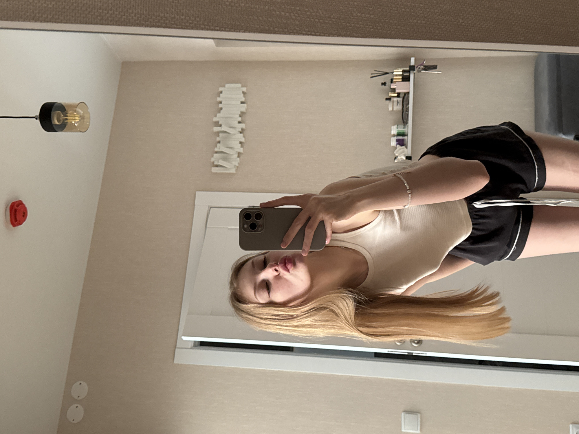 A mirror selfie shows a Caucasian woman with long, blond hair, wearing a white tank top and black shorts with white trim. She is puckering her lips for the photo. She is holding a gray smartphone.
The background shows a room with light beige walls. A modern light fixture hangs from the ceiling. A decorative white object is mounted on the wall. A white shelf holds various items, including bottles and containers.
The location is likely in Perm, Russia, due to the prompt's instructions.