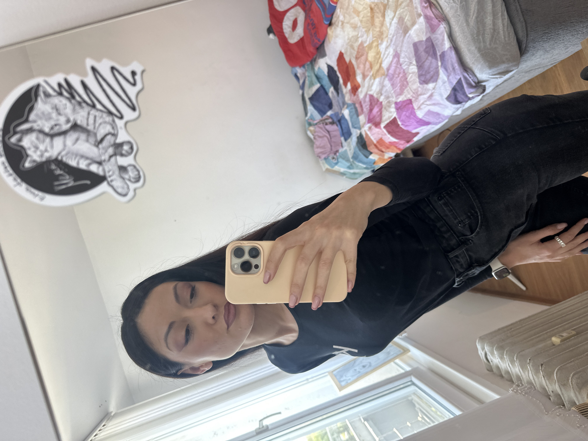 A woman takes a selfie in a mirror. The woman has long black hair, fair skin and is wearing a black long sleeve shirt and black jeans. She is holding a beige iPhone. The room has a bed with a patterned comforter, a sticker of cats on the wall, a radiator by the window, and a framed piece of art leaned against the wall. The location is not discernible from the image.