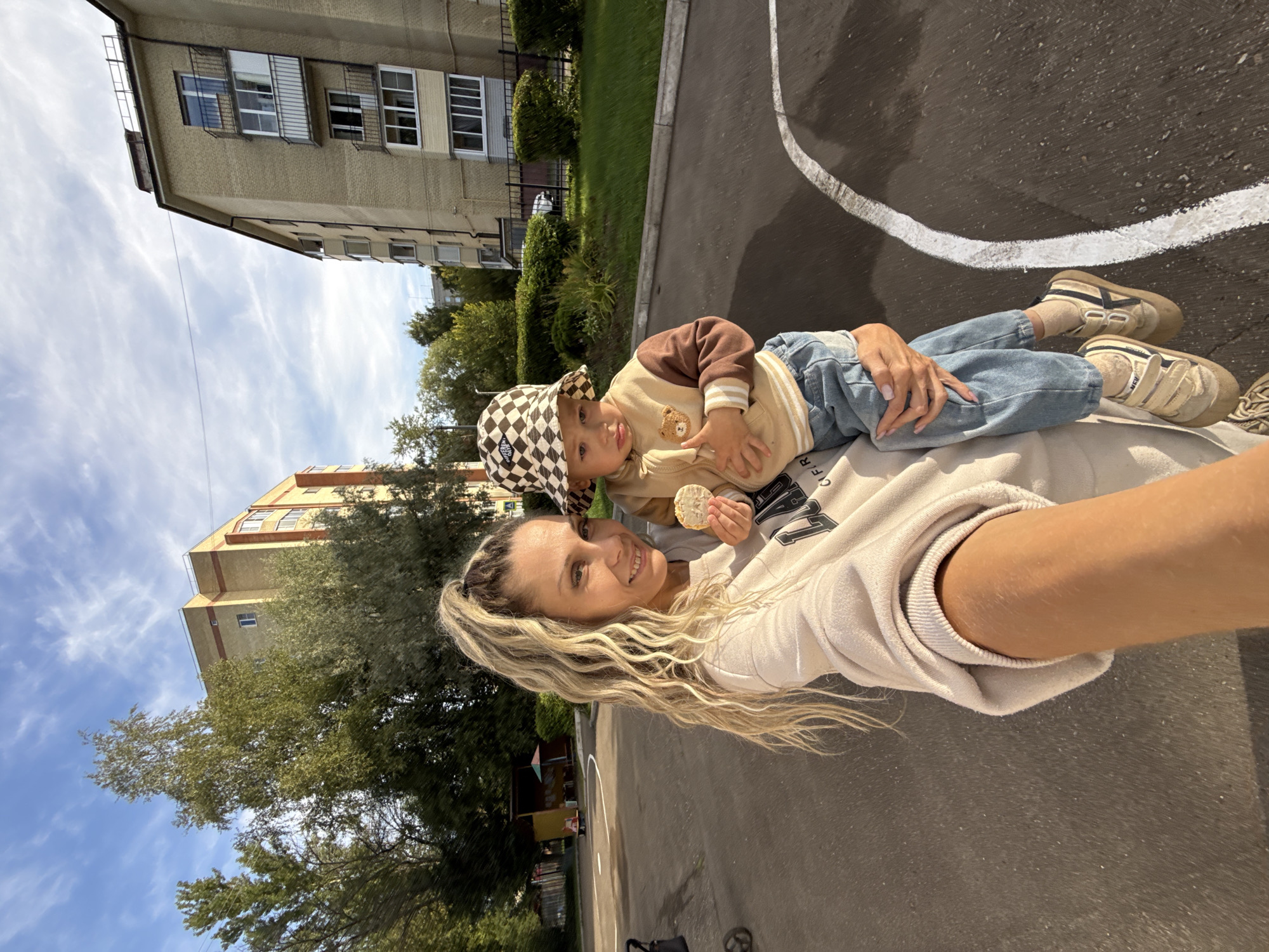 In Zhukovo, Russia, a woman with blonde, wavy, high-ponytail hair and fair skin is taking a selfie. She is wearing a beige sweatshirt. She is holding a young child who is wearing a checkered bucket hat and a beige and brown jacket with a bear patch, and blue jeans. The child is holding a small, light-colored biscuit in their hand. They are standing on a paved area, possibly a courtyard or playground, with white painted lines on the ground. In the background, there are apartment buildings, trees, and a children's play structure. The sky is blue with white clouds, and the sun is shining.