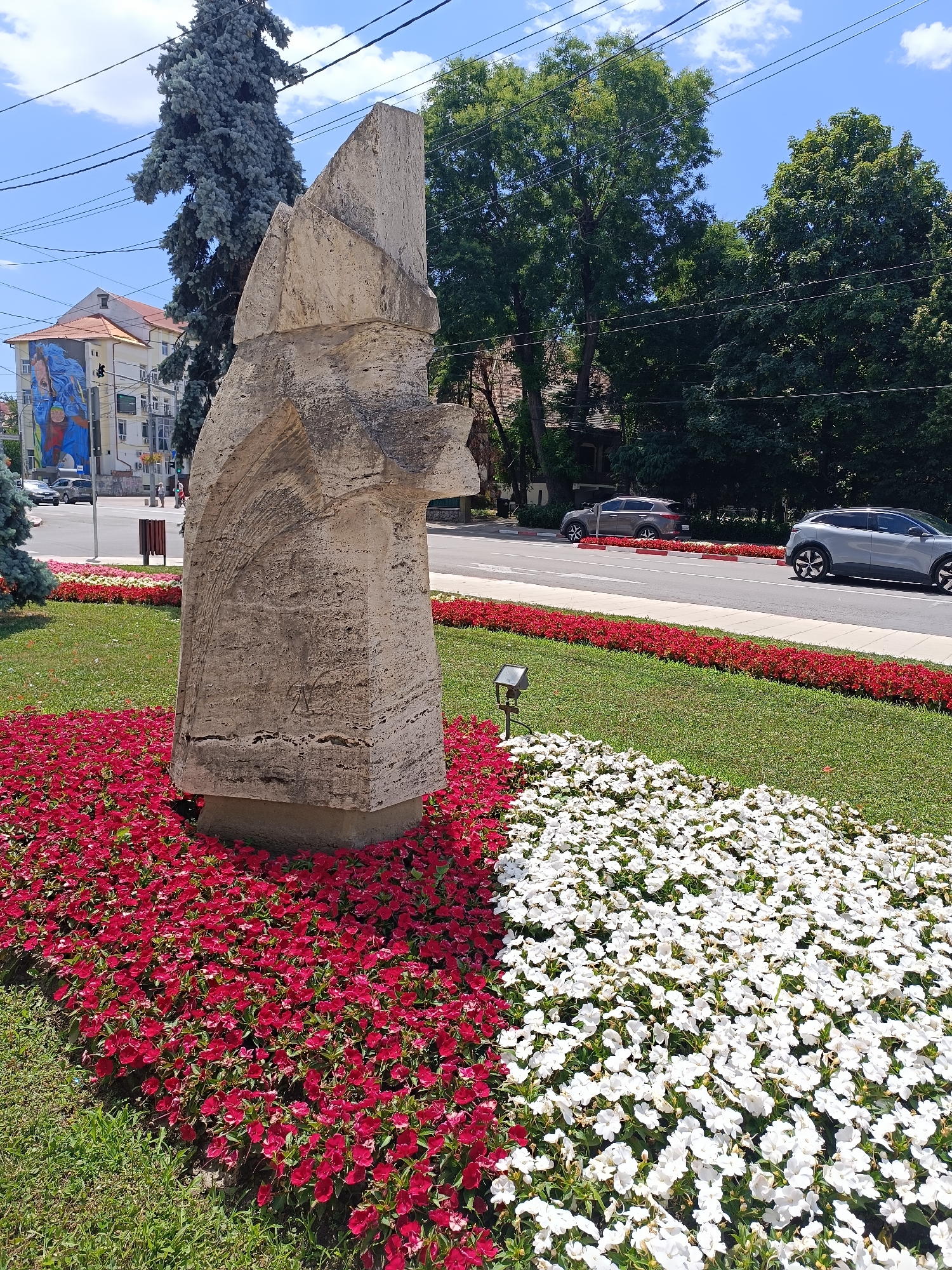 The image depicts a large, abstract stone sculpture situated in a landscaped area in Târgu Jiu, Romania. The sculpture is positioned in the foreground, surrounded by vibrant flower beds, with a row of red flowers to its left and white flowers to its right. Behind the sculpture, a street is visible with two cars driving on it. In the background, there are trees, buildings, and a clear blue sky with some clouds. A mural featuring a woman with blue hair is painted on the side of one of the buildings. The sculpture itself is made of rough-hewn, light-colored stone with distinct layers and textures. The overall scene suggests an urban environment with public art and urban greenery.