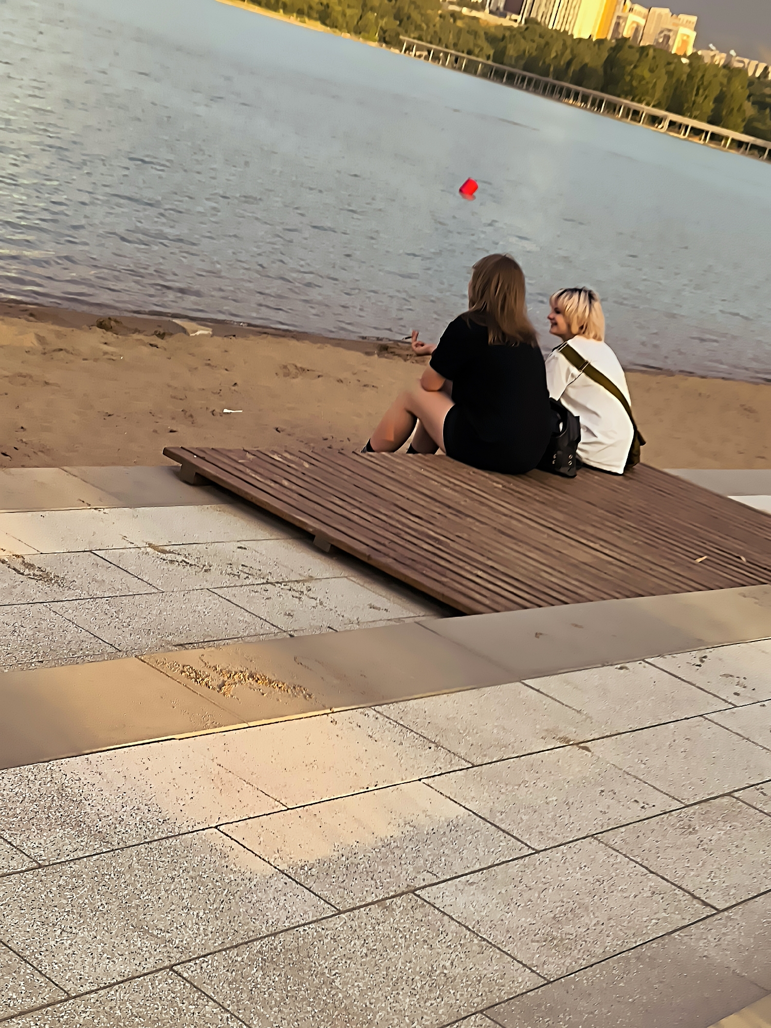 Two individuals are seated on a wooden platform near the shore of a body of water. The person on the left has long, light brown hair and is wearing a black t-shirt and black shorts. They are facing away from the camera and towards the water, with their left hand raised and appearing to be holding something small. The person on the right has short, blonde hair with pink streaks and is wearing a white t-shirt with a green strap across it. They are also facing away from the camera and towards the water, looking to their left and smiling. Both individuals have bags on the platform next to them. The water is calm and reflects the sky and distant cityscape. In the background, across the water, there is a line of trees and buildings, suggesting an urban environment. A red buoy is visible in the water to the left of the individuals. The foreground consists of a tiled promenade. The overall setting appears to be a recreational area by a lake or river within the city of Almaty, Kazakhstan.