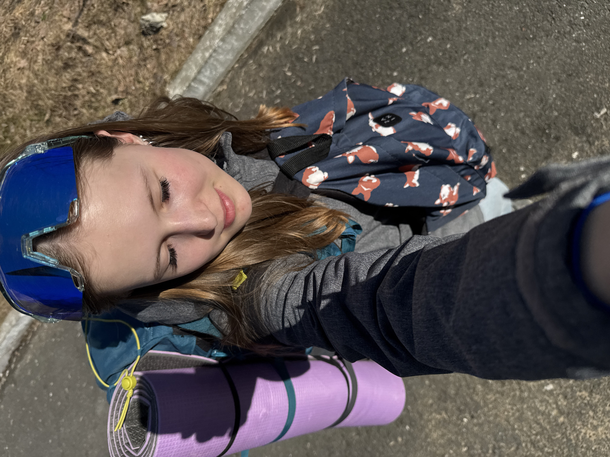 A young person is taking a selfie outdoors, likely in Berezniki, Russia. They are wearing ski goggles on their head and carrying a backpack with a fox pattern, as well as a purple and gray yoga mat strapped to the backpack. The background includes what appears to be pavement and some dry vegetation, suggesting it could be early spring. The person is smiling slightly with their eyes closed, looking towards the camera.