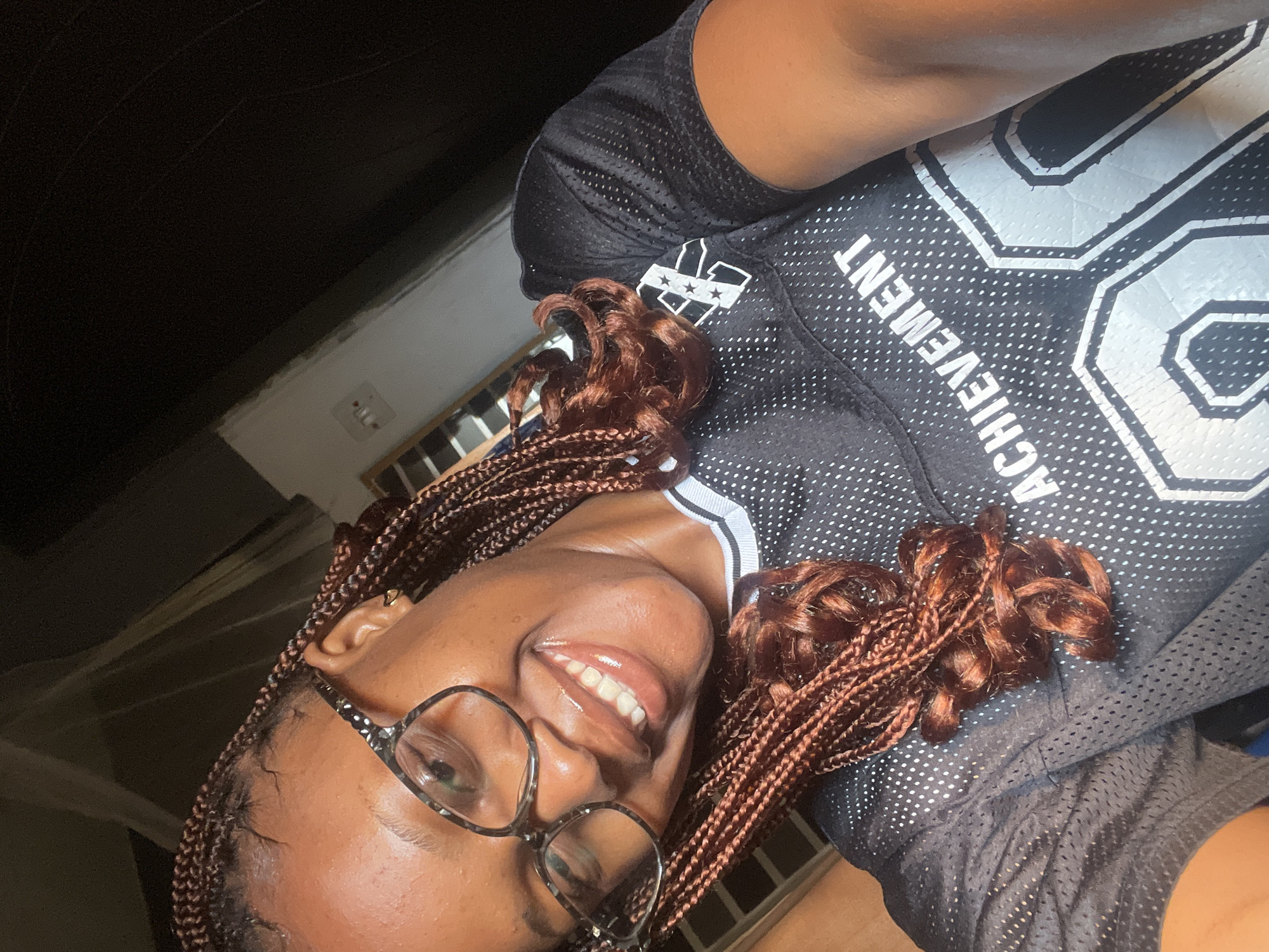 A young Black woman with braided hair and glasses smiles as she takes a selfie. She is wearing a black mesh jersey with white polka dots and white lettering that reads "ACHIEVEMENT 00". The background is dark, with a window and an electrical outlet visible on the right. The lighting suggests the photo was taken indoors, possibly in the evening. The image is cropped tightly on the woman's face and upper body. There is no specific indicator of location within Jos, Nigeria, but the setting appears to be residential.