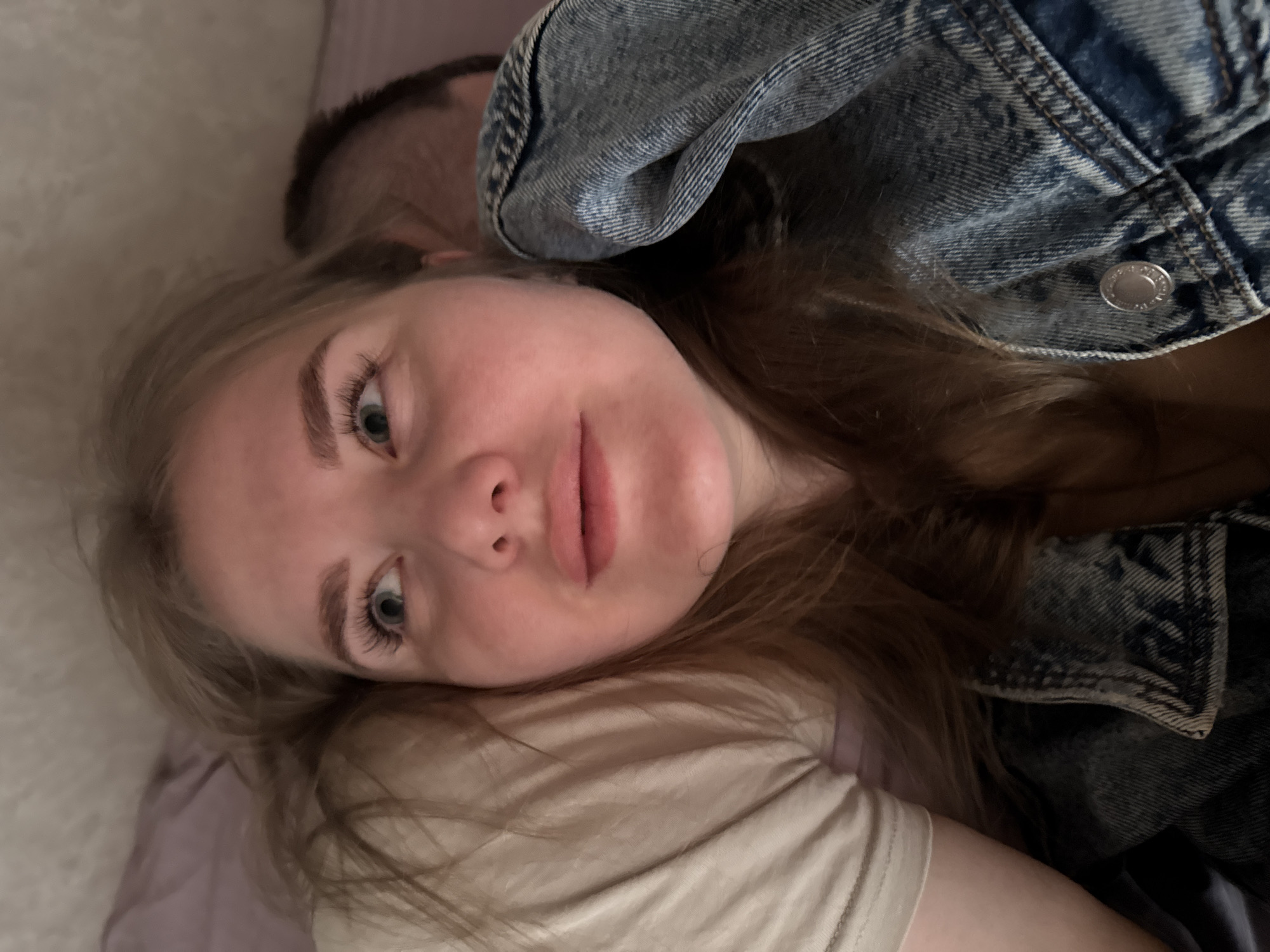A close-up, angled view of a fair-skinned woman with light brown hair and blue eyes lying down. She is wearing a beige t-shirt and a blue denim jacket is partially draped over her shoulder. Part of a man's head is visible in the background. The setting seems to be an indoor, casual environment. The location in the city of Gornyi (Dvurechenskoe S/P), Russia is not evident from the image.