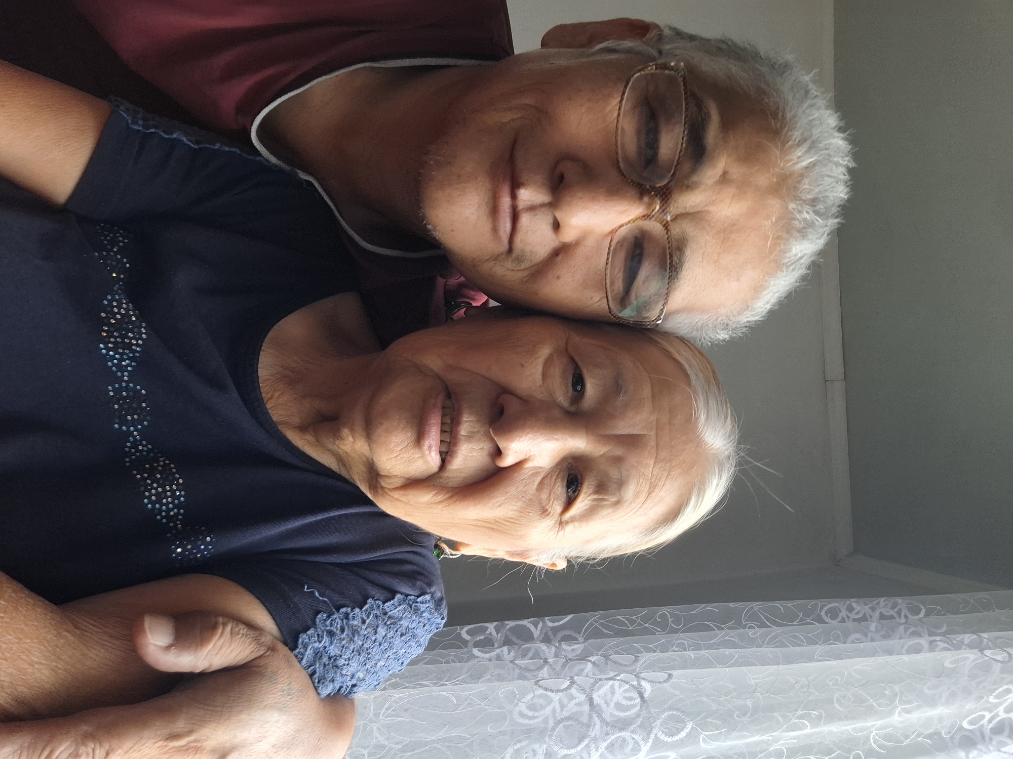 A waist-up, eye-level, outdoor selfie shows two individuals, a man and an elderly woman, positioned closely together. The man, on the left, wears glasses and a maroon t-shirt with a white trim. He has graying hair and appears to be smiling subtly. The woman, on the right, has white hair and wears a dark blue t-shirt with blue lace detailing on the shoulder and a band of rhinestones across the chest. She has a green earring in her visible ear and is looking towards the camera. The man's arm is around the woman's shoulders, and her arm is resting on his. They are indoors, as indicated by the presence of a light-colored wall and a white lace curtain visible on the right side of the frame, suggesting a domestic setting. The lighting suggests it is daytime, with natural light coming from the window. The scene appears to be in Almaty, Kazakhstan, given the metadata.