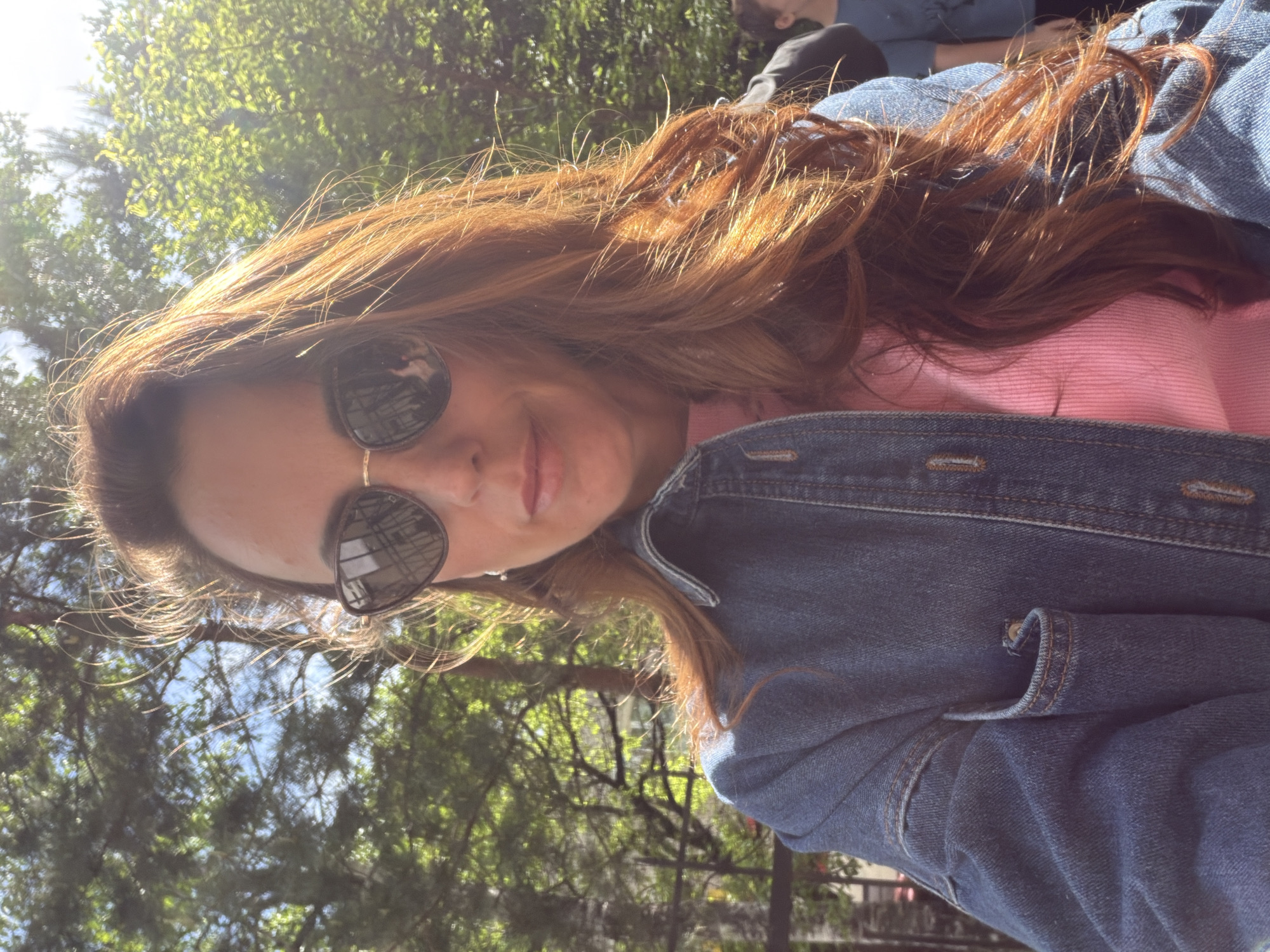 A woman with long, reddish-brown hair is posing for a selfie outdoors. She is wearing dark sunglasses, a pink top, and a blue denim jacket. The background shows trees and foliage, suggesting a park or wooded area. Other people can be seen in the background, though they are out of focus. The picture has a sunny, outdoor feel. It is not possible to determine that the picture was taken in Birobidzhan, Russia, as there is nothing in the image to indicate the location.